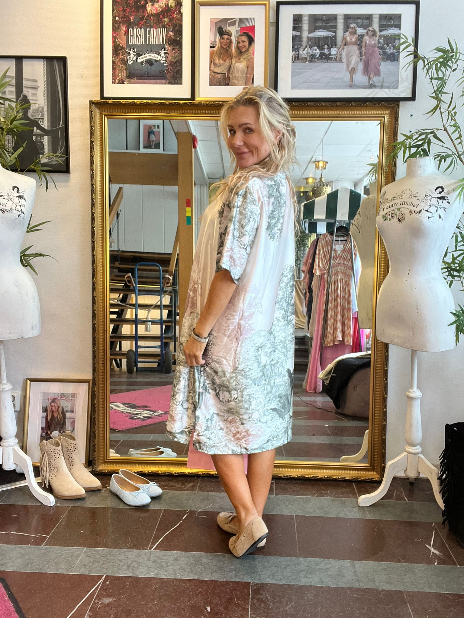 Blond kvinna står framför en stor guldramsspegel och tittar leende över axeln medan hon provar en ljus, mönstrad kimono i en butik. Runt henne syns skyltdockor, skor på golvet och tavlor, inklusive texten “CASA FANNY”.