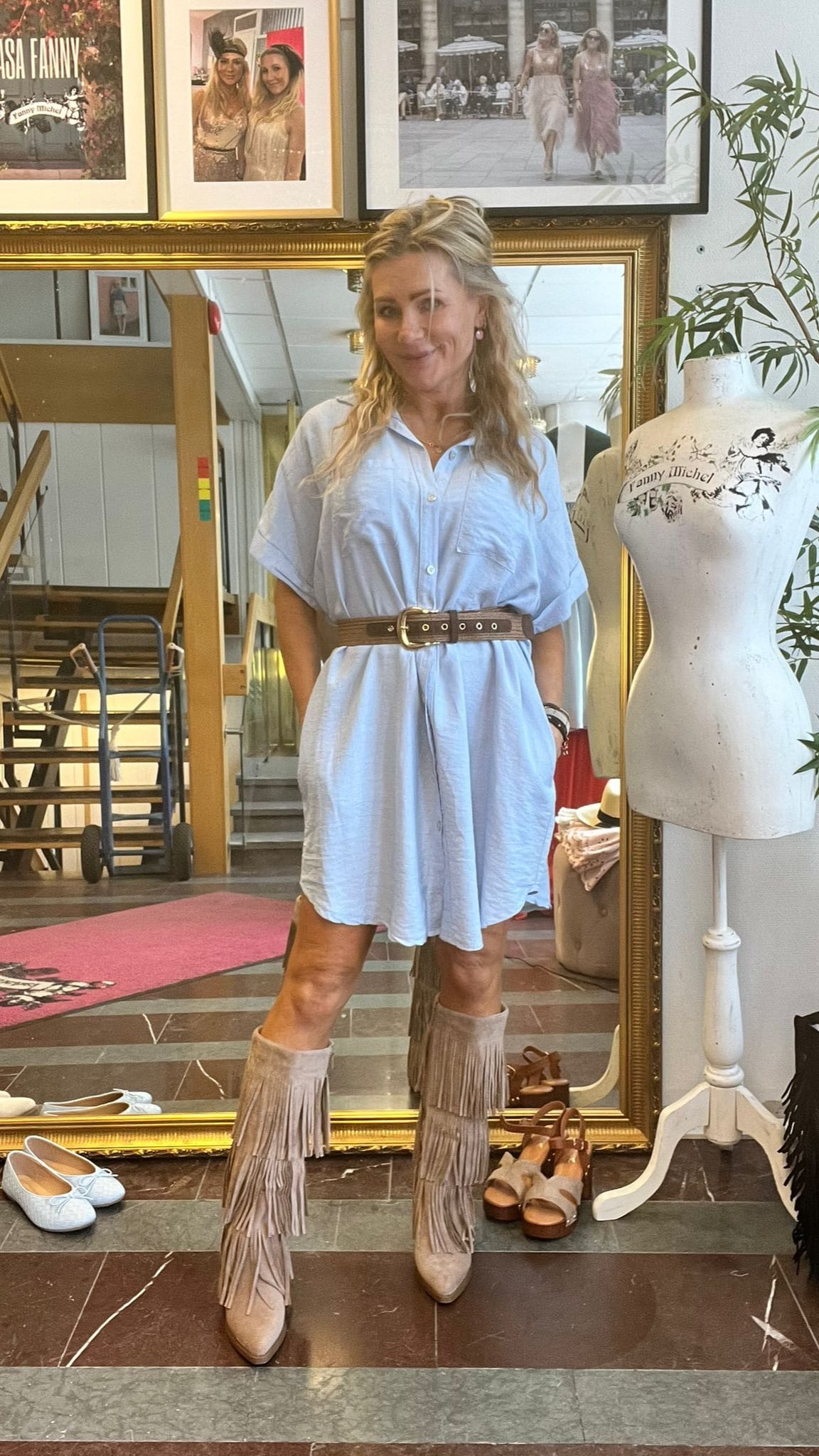 Blond kvinna står och poserar framför en stor guldramsspegel i en butik, med händerna i fickorna. Hon bär ljusblå skjortklänning med brunt bälte och fransade stövlar. Intill syns provdocka med texten ”Fanny Michel”.