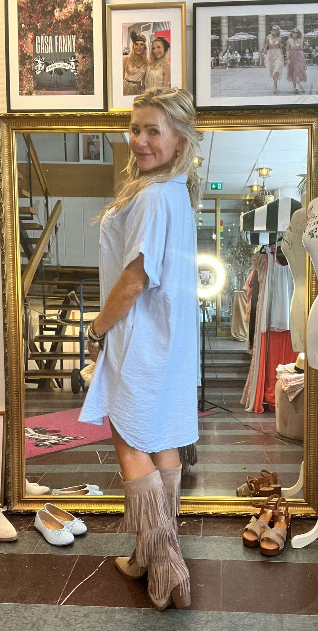 Blond kvinna poserar i profil framför en stor spegel i en klädbutik, klädd i ljus tunika och fransstövlar. Skor ligger på golvet och klänningar hänger bredvid. Inramade foton ovanför, text: ”CASA FANNY”.
