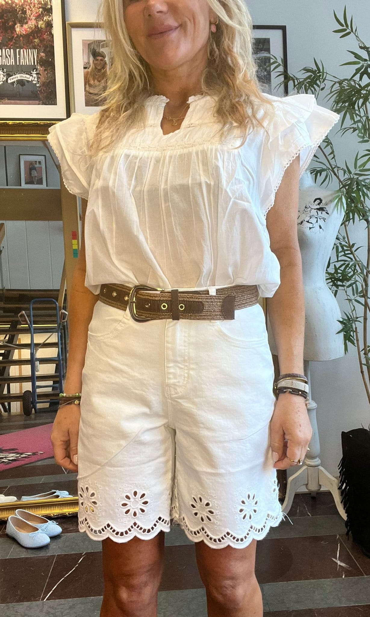 Kvinna står inomhus och visar sin sommaroutfit: vit blus med volangärmar och vita broderie anglaise-shorts, markerat med brett brunt bälte. I bakgrunden syns inramade tavlor och en stor växt; ljusa skor på golvet.