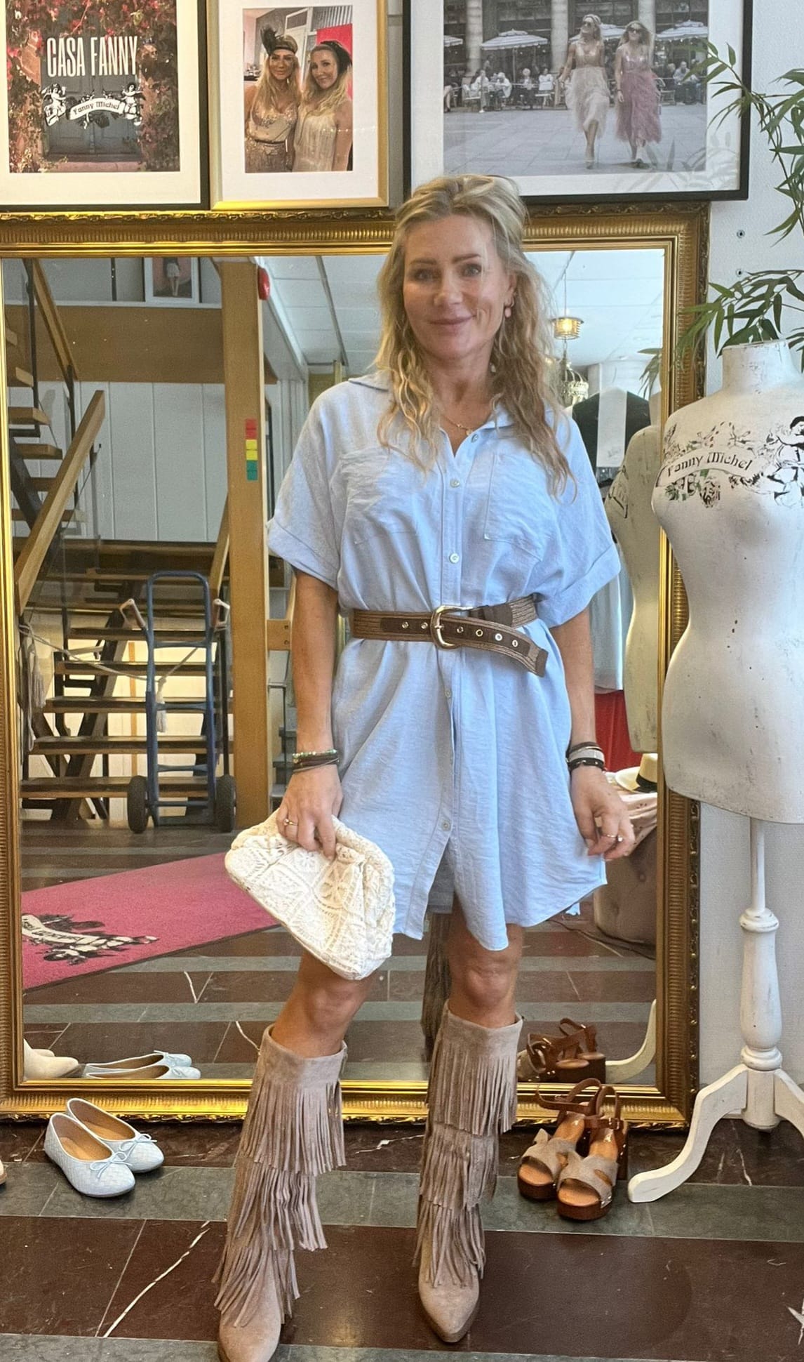 Kvinna står och poserar framför en stor guldramad spegel i en butik, klädd i ljusblå skjortklänning med brett brunt bälte, fransade stövlar och vit clutch. På väggen syns texten “CASA FANNY”.