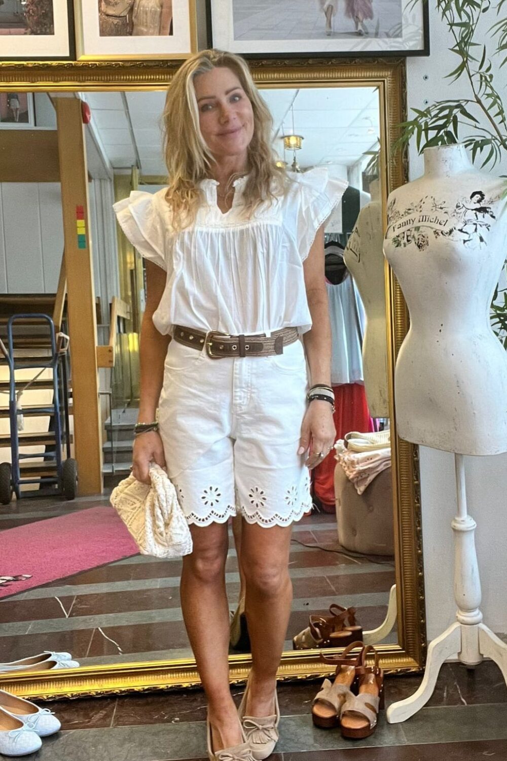 Leende kvinna står framför en stor guldramsspegel och visar en somrig outfit: vit volangblus, vita broderade shorts, brunt bälte och beige loafers, med en liten flätad väska i handen i en butik med provdocka och skor.
