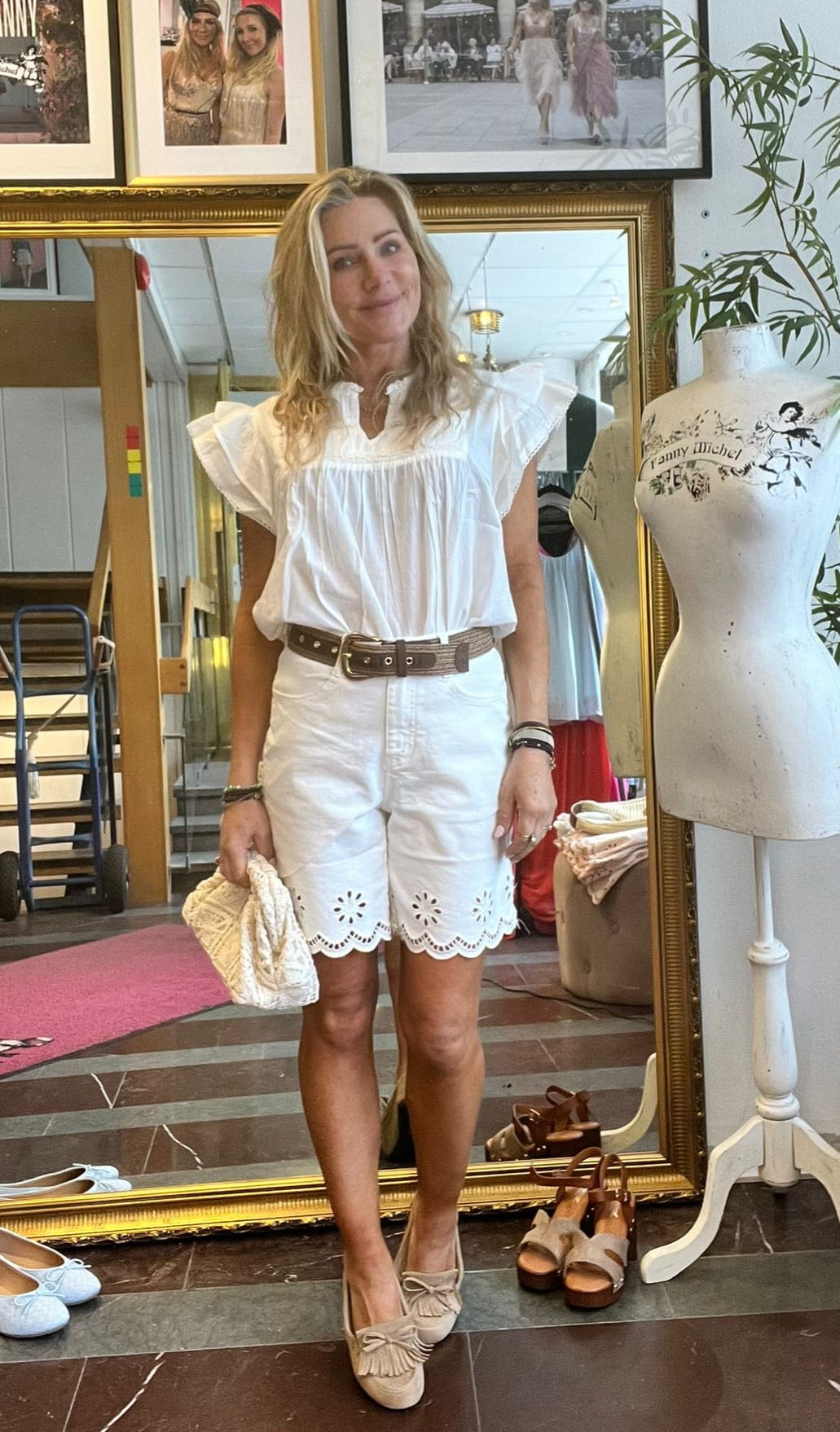 Leende kvinna står framför en stor guldramsspegel och visar en somrig outfit: vit volangblus, vita broderade shorts, brunt bälte och beige loafers, med en liten flätad väska i handen i en butik med provdocka och skor.