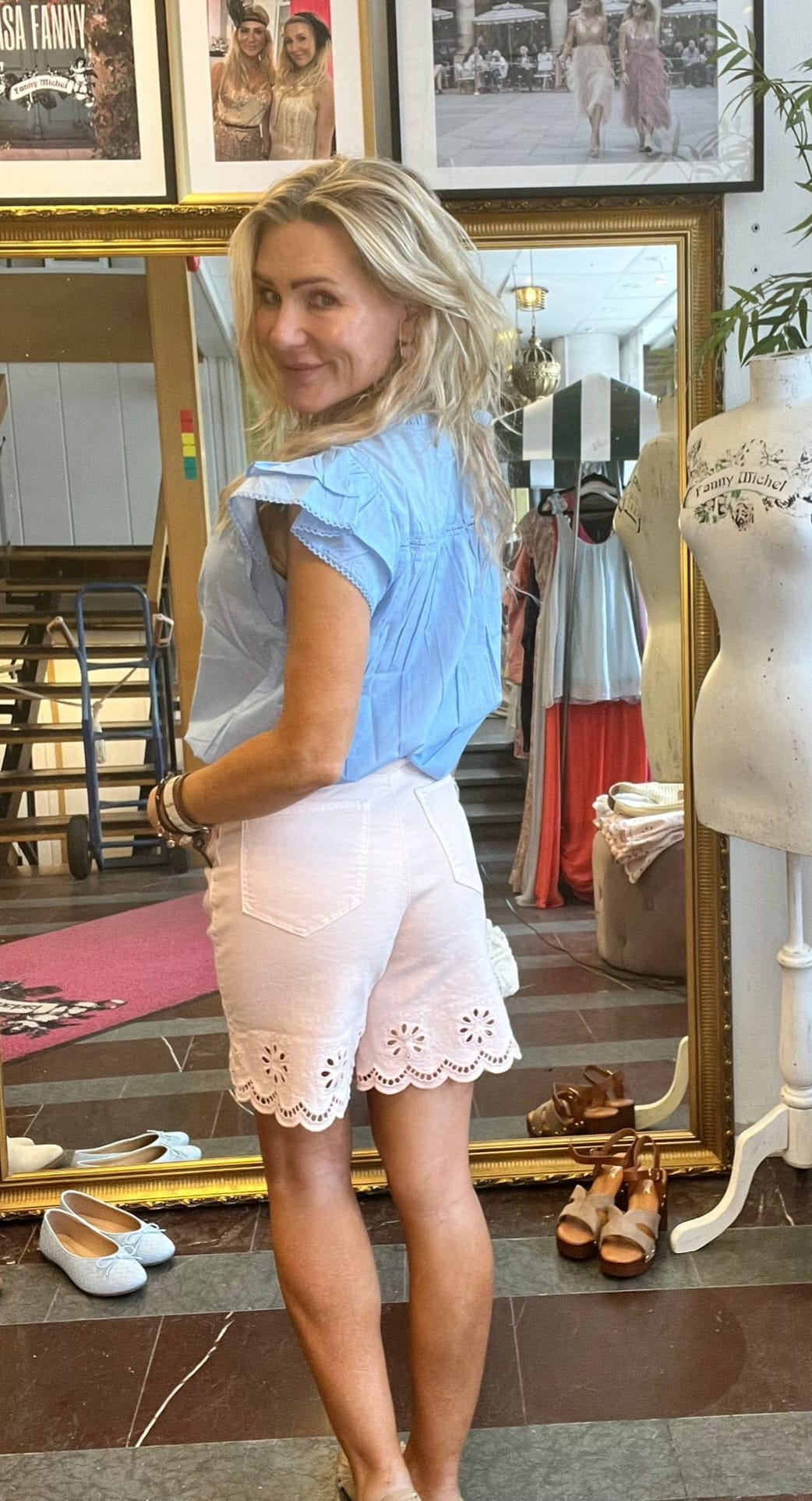 Blond kvinna provar kläder och vänder sig leende mot kameran, iklädd ljusblå blus och vita shorts med broderad kant. Hon står framför en stor guldramsspegel i en butik med skor, klädställning och provdocka. Text: “Fanny Michel”.