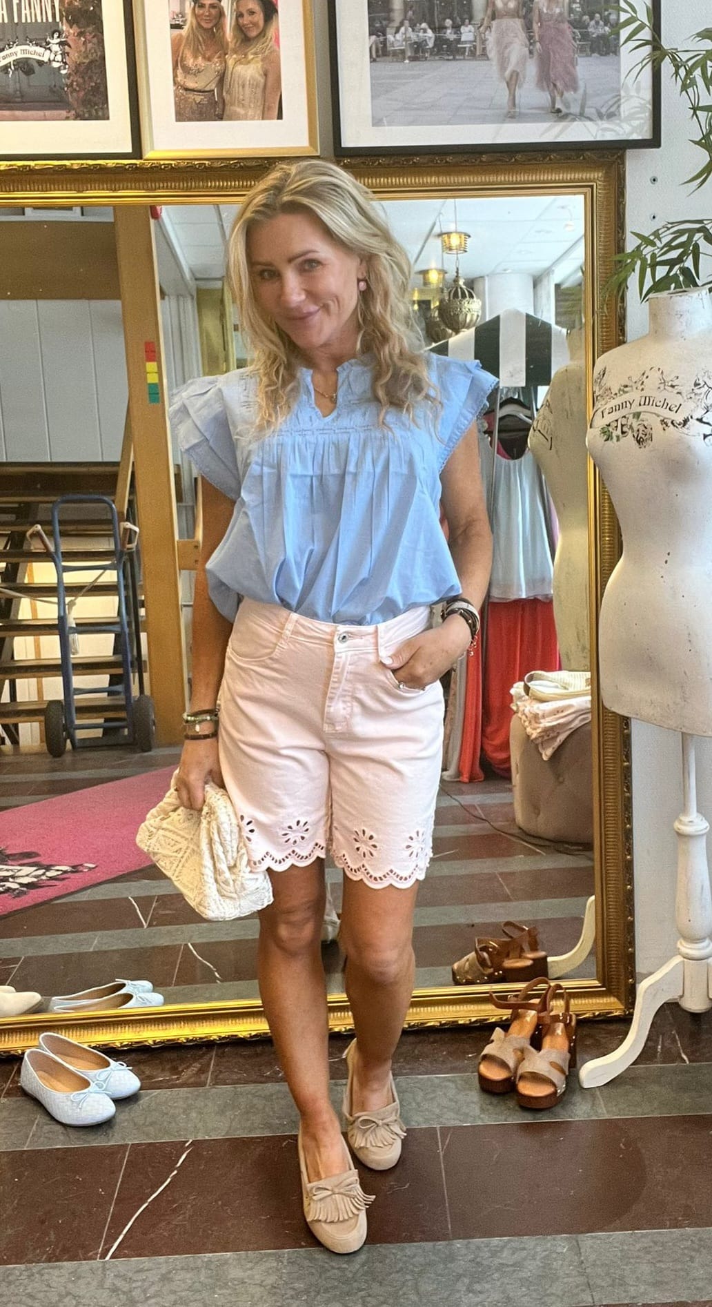 Blond kvinna poserar framför en stor guldramsspegel i en butik, med handen i fickan och en stickad väska. Hon bär ljusblå blus, ljusrosa shorts med brodyr och beige loafers; skor och skyltdocka syns. Text: “Fanny Michel”.