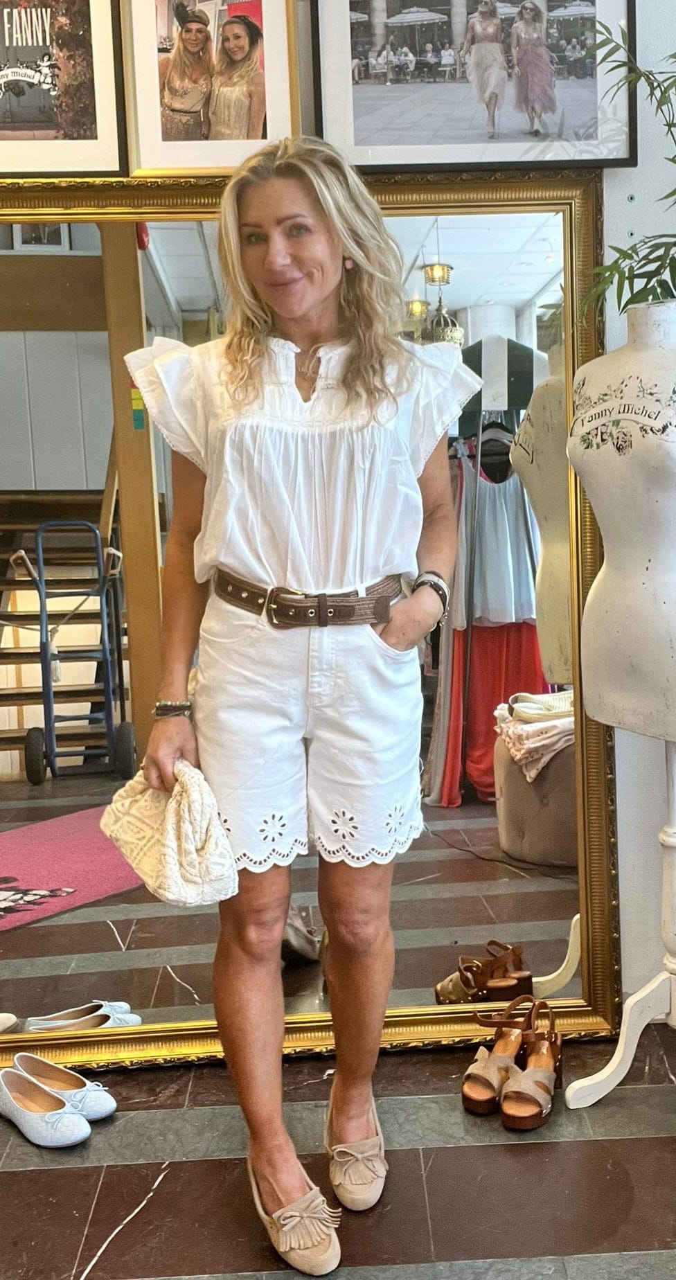 Kvinna står och posar framför en stor guldramad spegel i en klädbutik, med handen i fickan och en quiltad väska i andra. Hon bär vit blus och vita broderade shorts; skor ligger på golvet. Text: “FANNY”.