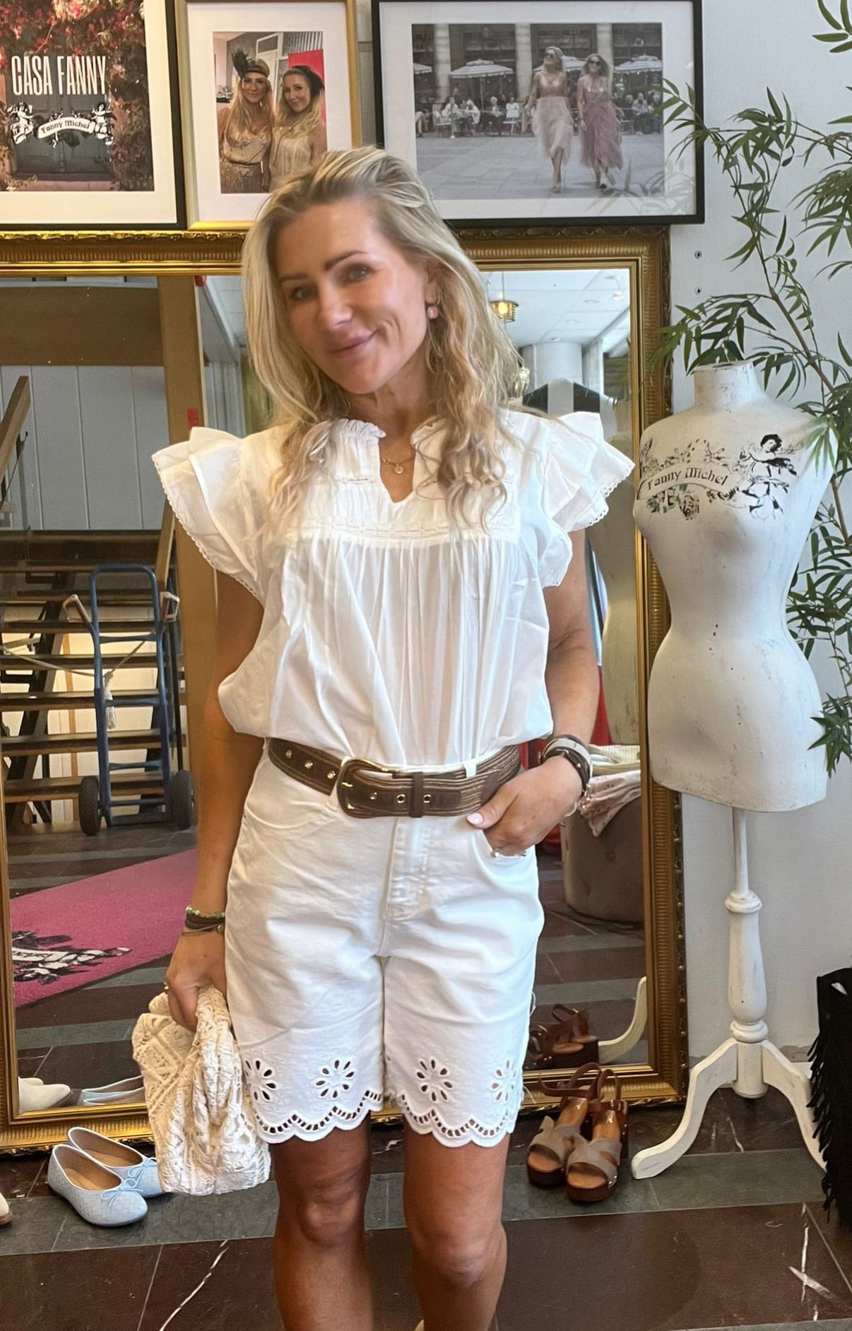 Leende kvinna i vit blus och vita shorts med bredt brunt bälte poserar med handen i fickan och håller en beige väska. Bakom syns en stor spegel, skor på golvet och ramar med texten “CASA FANNY” och “Fanny Michel”.