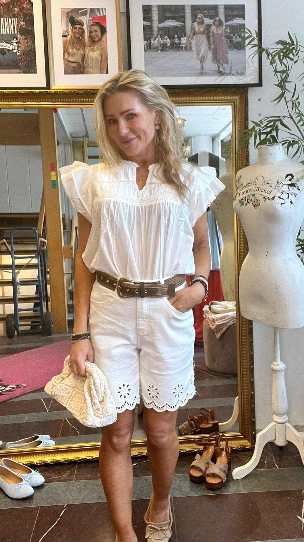 Leende kvinna poserar framför en stor guldramsspegel i en butik, klädd i vit volangblus och vita broderade shorts med brunt bälte, håller en ljus flätad väska. Bredvid står provdocka med texten “Jenny Michel” och skor på golvet.