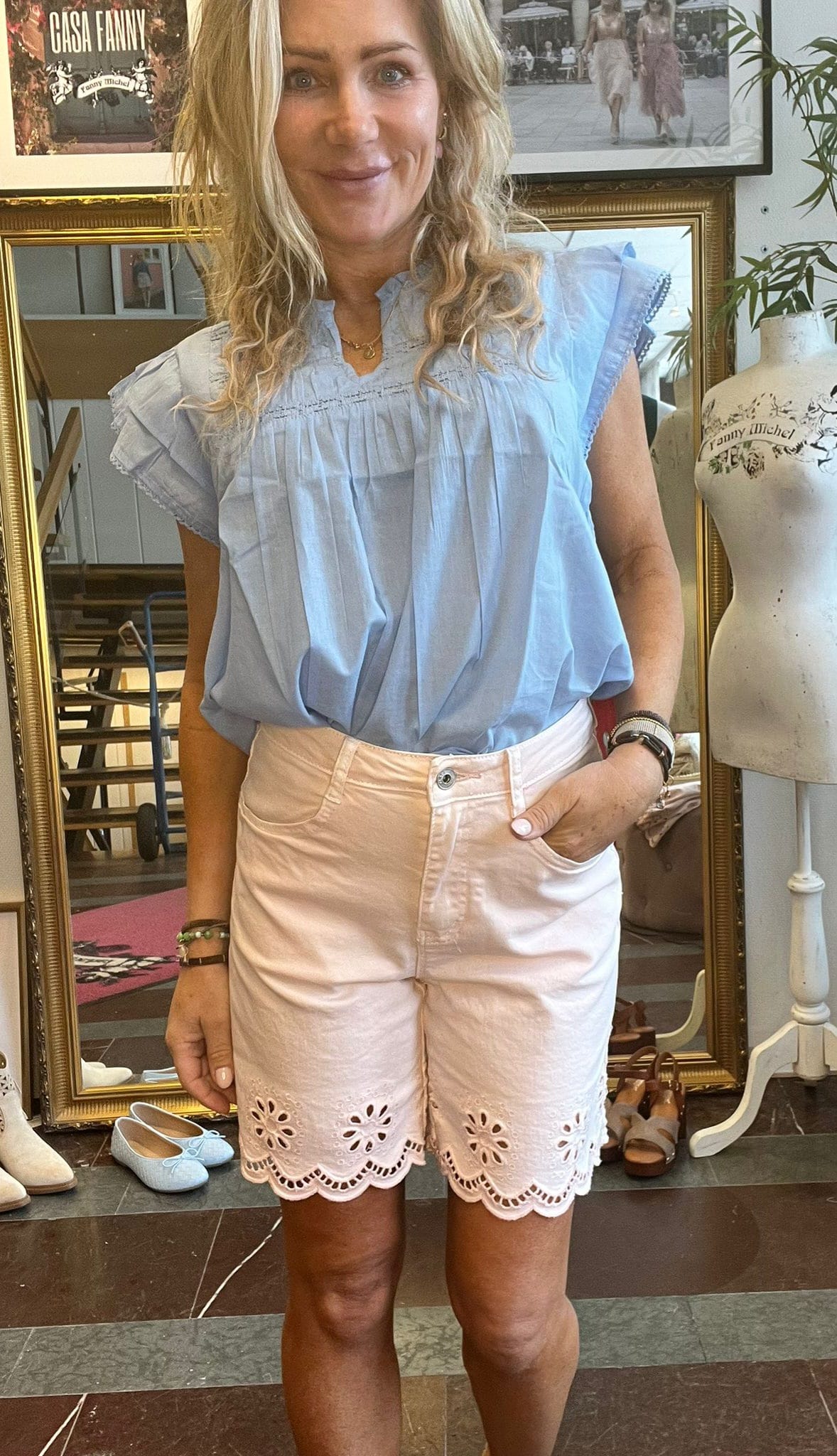 Kvinna står och ler med en hand i fickan, klädd i ljusblå volangtopp och ljusrosa shorts med broderad kant. Hon står framför en spegel i en butik/ateljé med skor och provdocka; text syns: ”CASA FANNY”, ”Fanny Michel”.