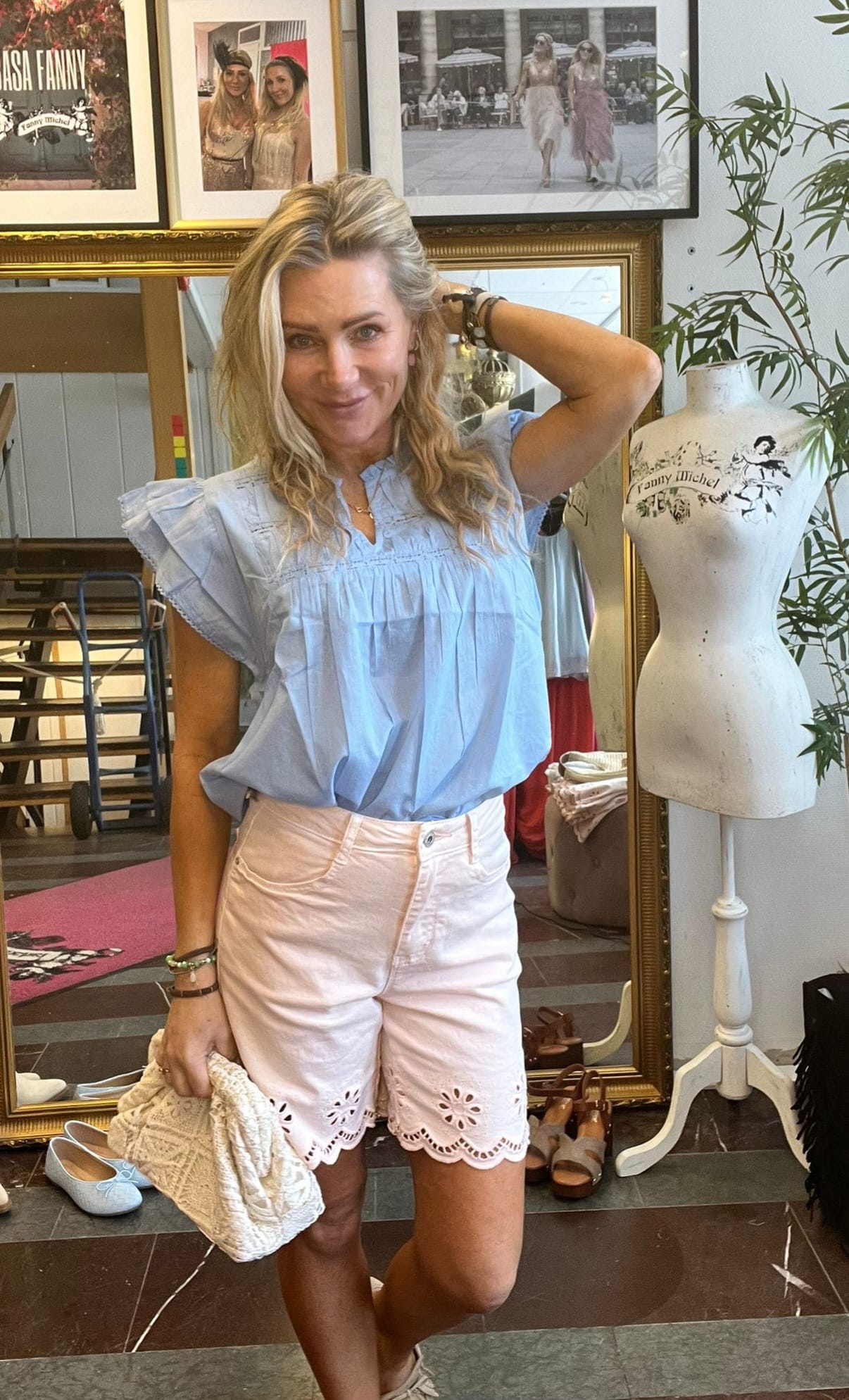Leende kvinna med blont vågigt hår poserar inomhus, med ena handen bakom huvudet och en stickad väska i den andra. Hon bär ljusblå blus och rosa shorts; bakom syns spegel, skor, provdocka och tavlor med texten “Fanny Michel”.