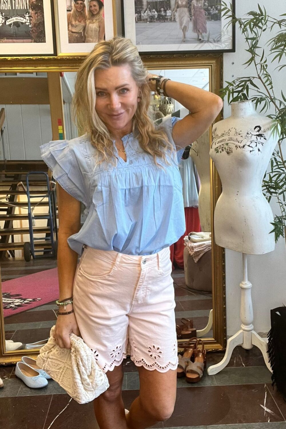 Blond kvinna poserar och ler med ena handen bakom huvudet och en stickad väska i den andra, klädd i ljusblå blus och ljusrosa shorts. Hon står i en butik med spegel, skor och skyltdocka med texten ”Fanny Michel”.