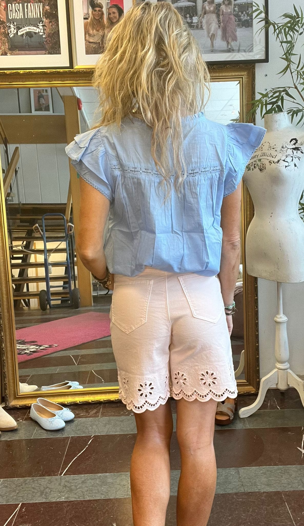 Kvinna med blont, vågigt hår står med ryggen mot kameran och tittar i en stor guldramad spegel i en butik. Hon bär ljusblus och vita shorts; bredvid syns provdocka och skor. Text: “CASA FANNY”.