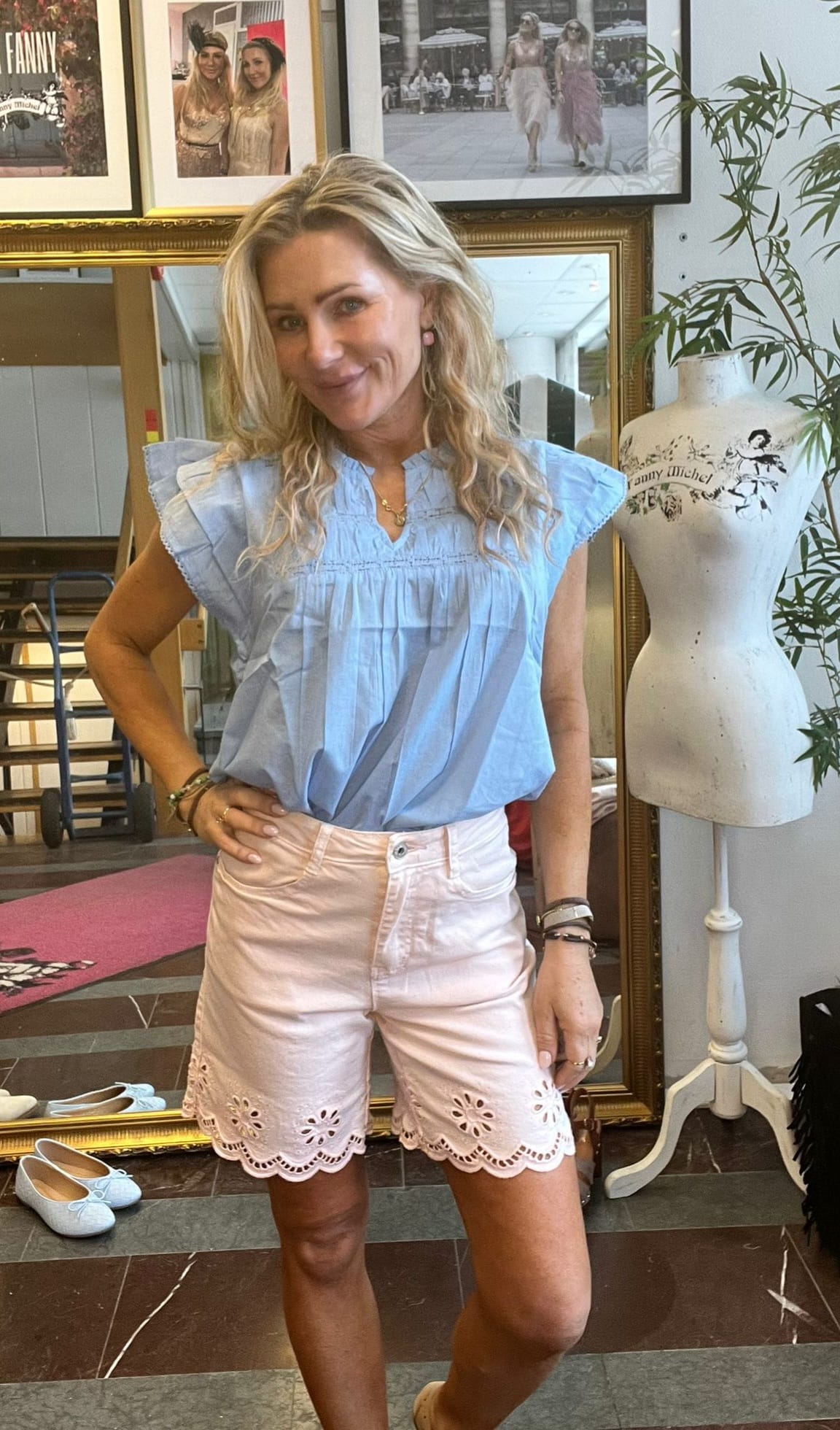 Leende kvinna står med handen på höften i en klädbutik eller studio, klädd i ljusblå blus och rosa shorts med brodyrkant. Bakom syns stor spegel, inramade foton och provdocka med texten “Fanny Michel”.