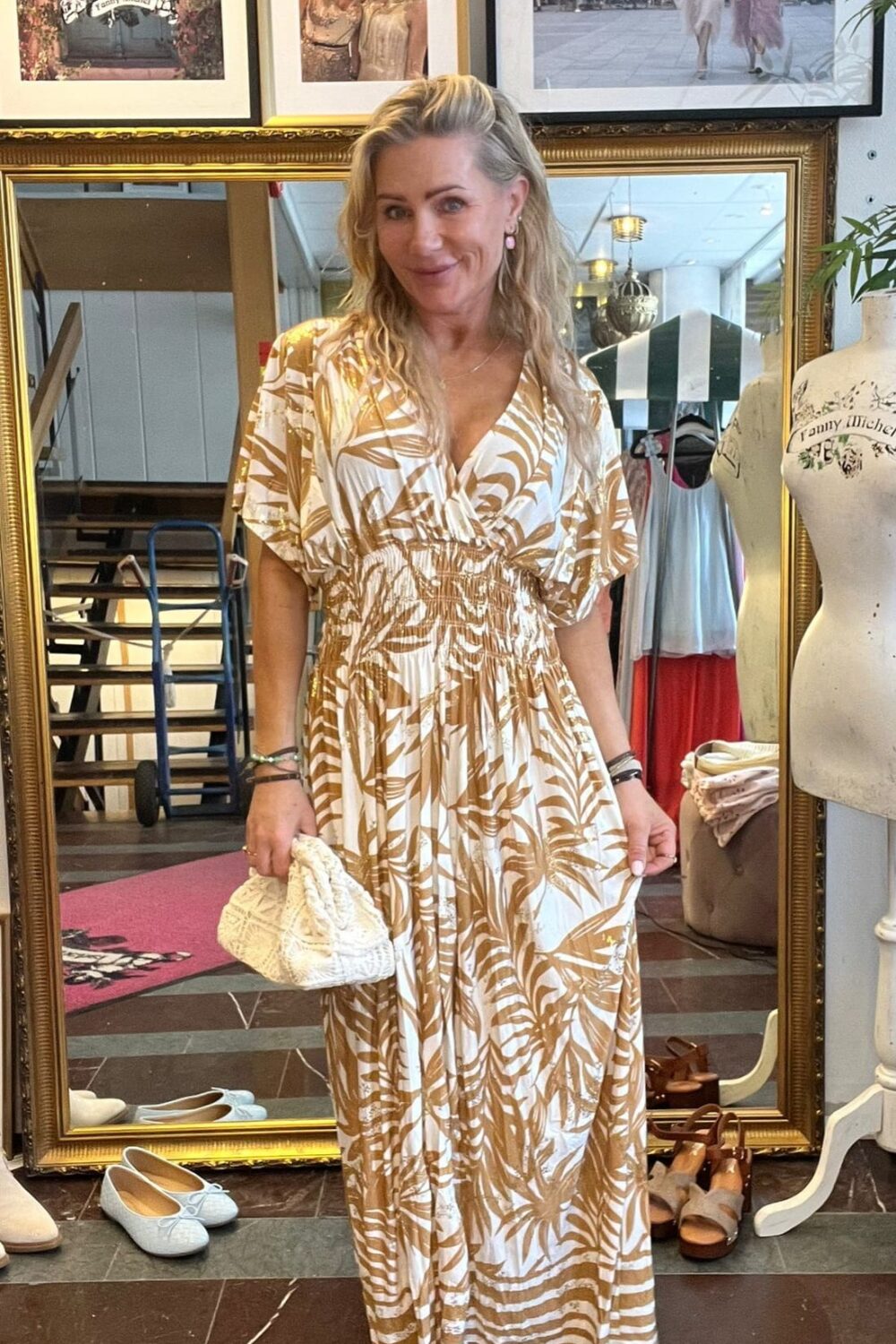 Leende kvinna står i en butik och håller ut sidorna på en beigevit, mönstrad maxiklänning samt en liten vit clutch. Bakom syns spegel, skyltdocka, skor på golvet och inramade foton med texten “CASA FANNY” och “Fanny Michel”.