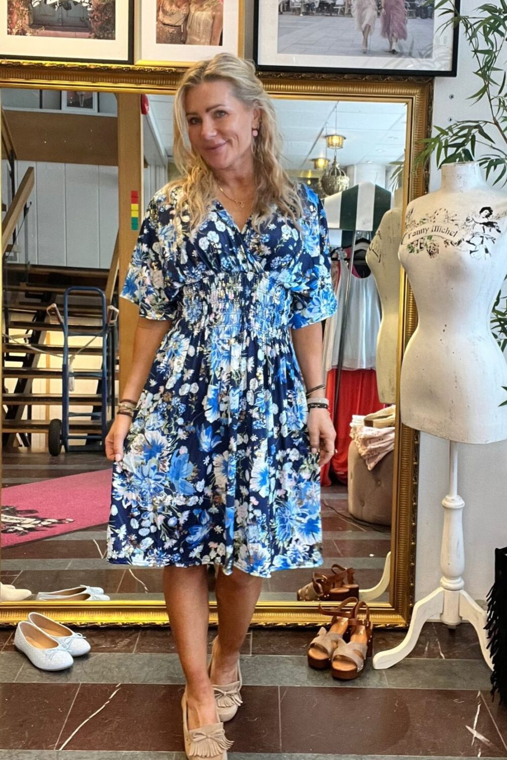 Leende kvinna står framför en stor guldramsspegel och visar upp en blå blommig klänning och beige loafers i en butik. Runt henne syns skor på golvet, skyltdocka med texten ”Fanny Michel” och inramade foton.