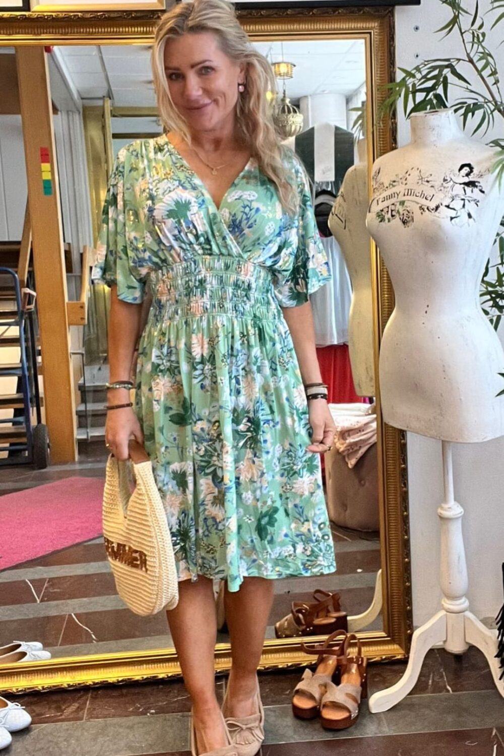 Blond kvinna poserar och ler i en butik, klädd i grön blommig klänning och ljusa loafers, med en halmhandväska i handen. Bredvid står en provdocka med texten ”Lilly Michel”, spegel och inramade foton i bakgrunden.