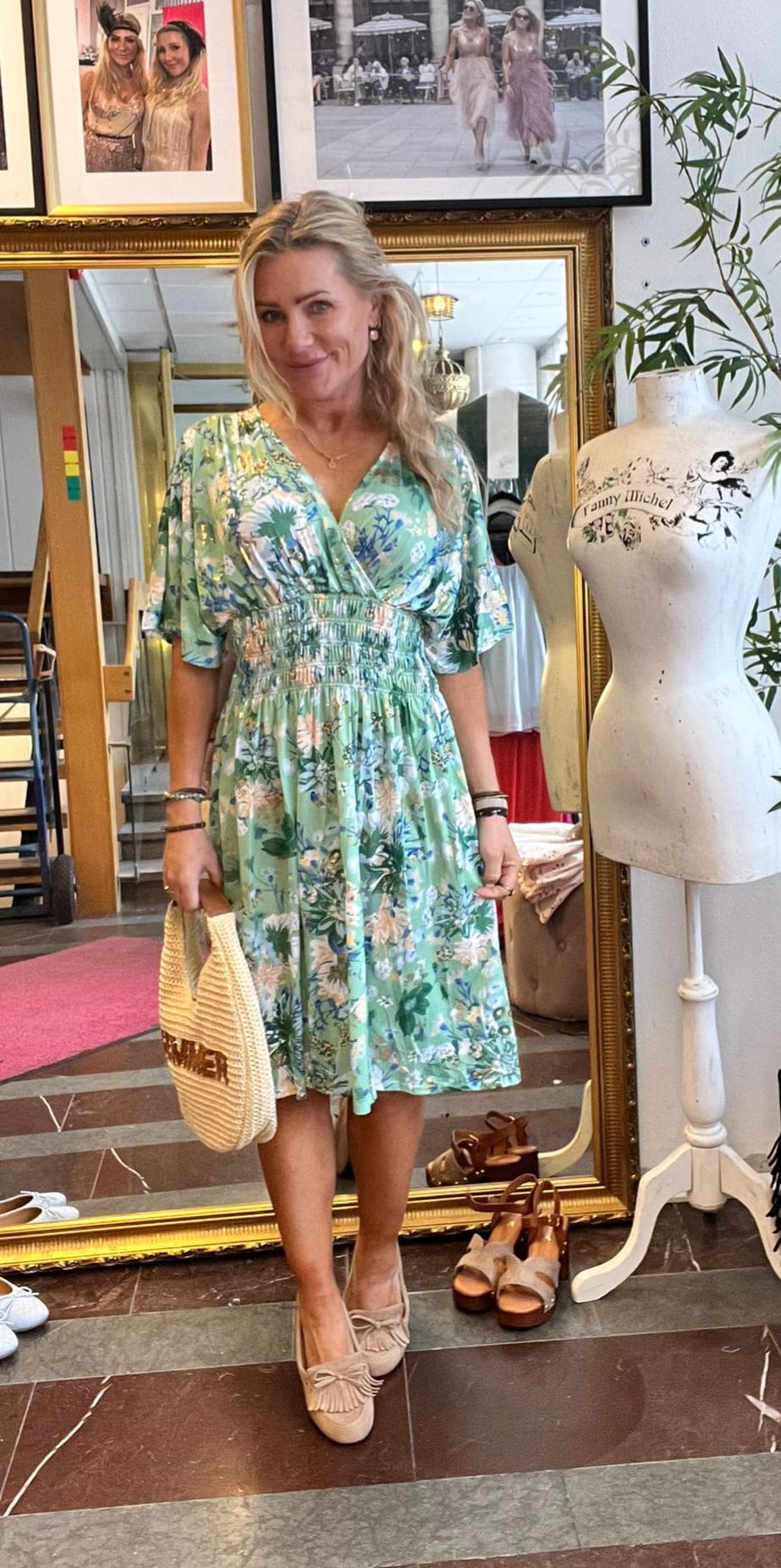 Blond kvinna poserar och ler i en butik, klädd i grön blommig klänning och ljusa loafers, med en halmhandväska i handen. Bredvid står en provdocka med texten ”Lilly Michel”, spegel och inramade foton i bakgrunden.