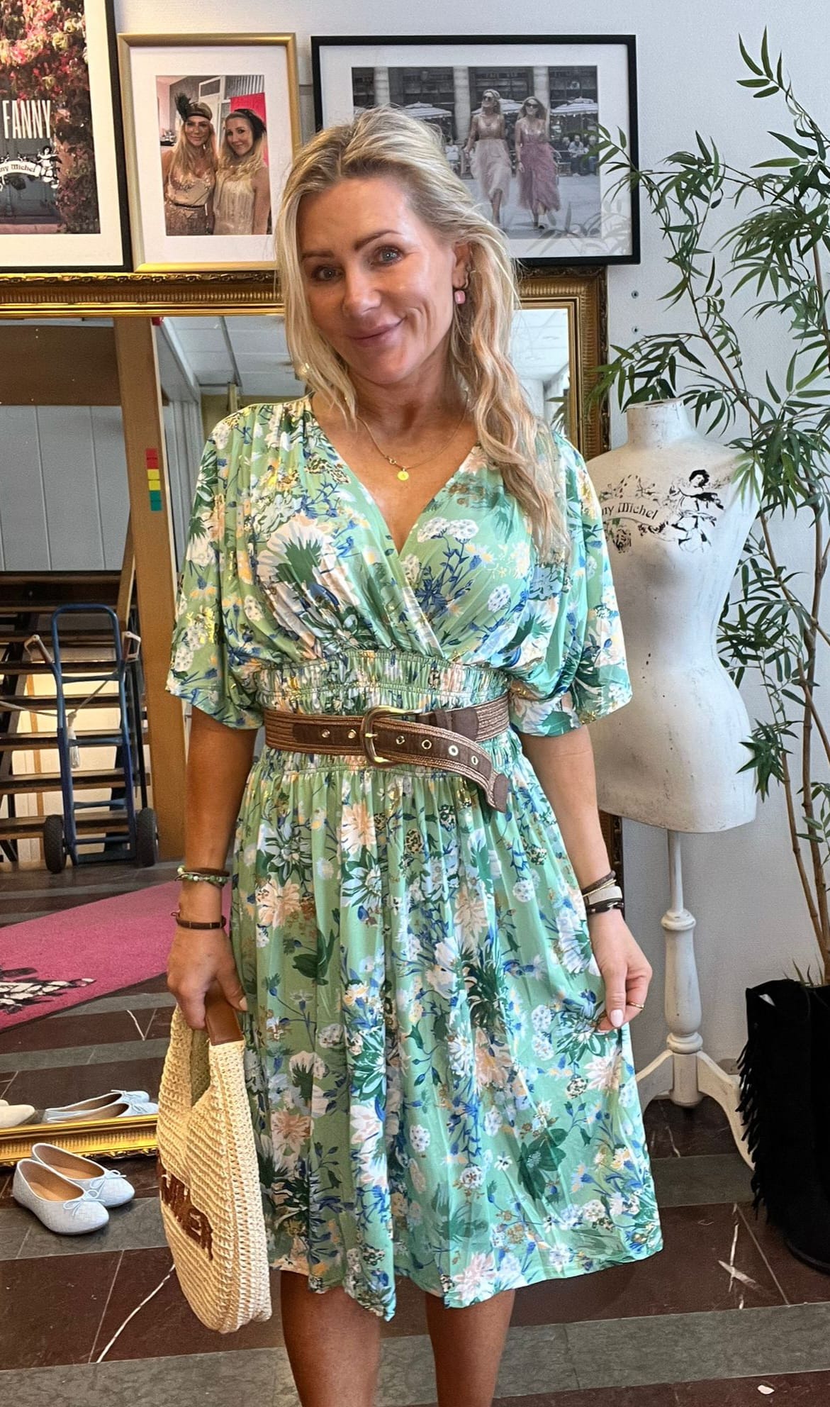 Blond kvinna står och ler mot kameran i en butik eller ateljé, iklädd grön blommig klänning med brett brunt bälte och halmväskan i handen; bakom syns inramade foton, spegel, provdocka och växt.