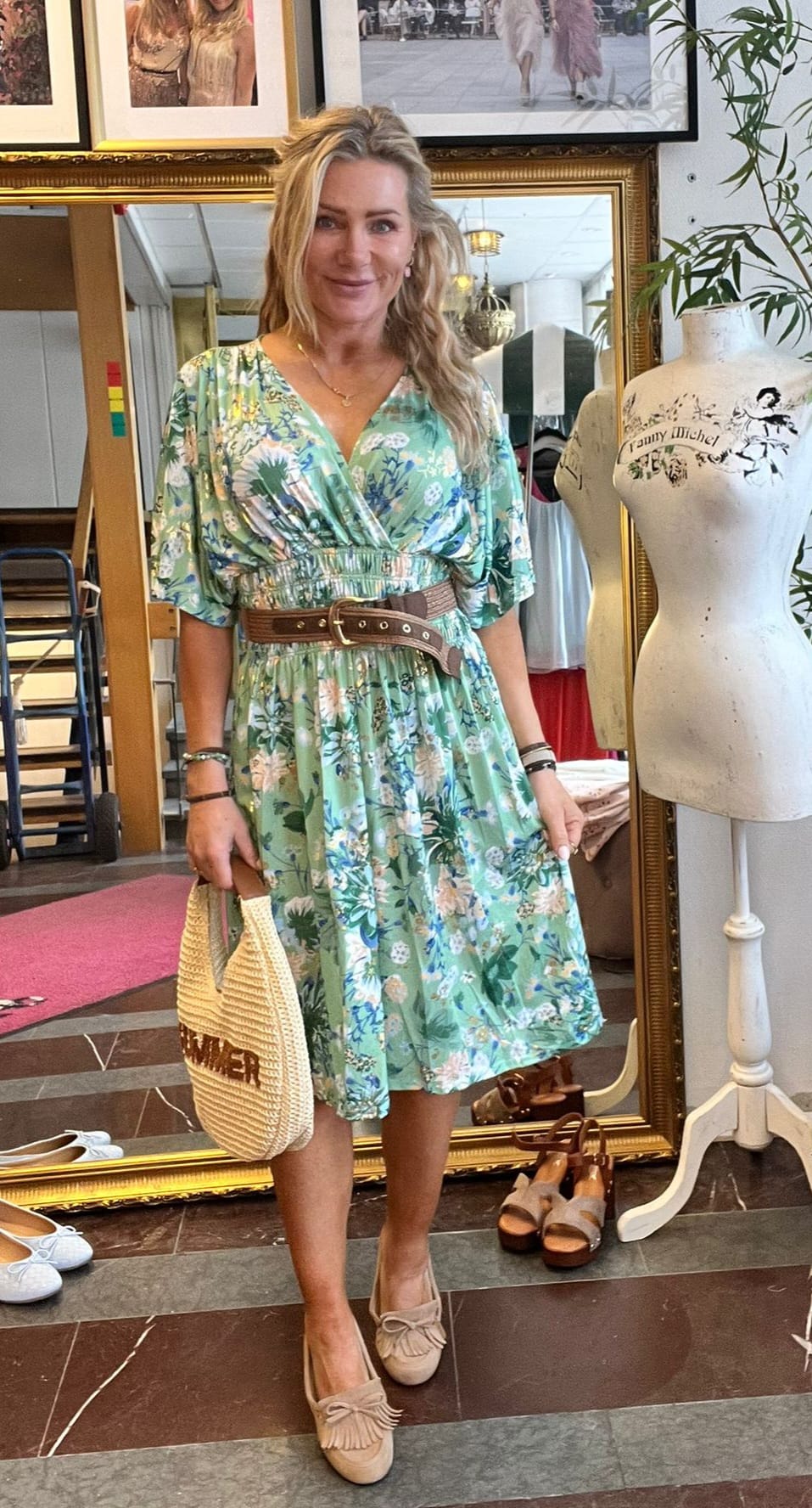 Leende kvinna står i en butik och visar en grön, blommig klänning med brunt bälte, håller en stråväska med texten ”SUMMER”. Bredvid syns provdocka med texten ”Fanny Michel”, spegel, skor och tavlor.