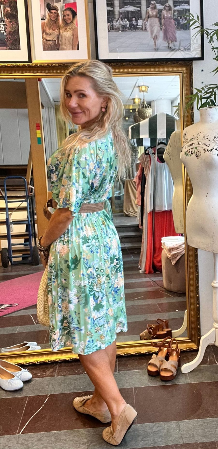 Blond kvinna i grön, blommig klänning står framför en stor guldramsspegel och tittar leende över axeln. Hon håller en stråväska i en klädbutik med provdocka märkt “Family Michel”, skor på golvet och klädställning.