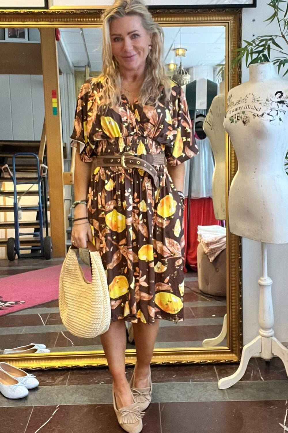 Blond kvinna står och ler framför en stor guldramsspegel i en butik, iklädd brun klänning med fruktmönster och skärp, håller en ljus stråväska. Skyltdocka bredvid; text på den: “Fanny Michel”.