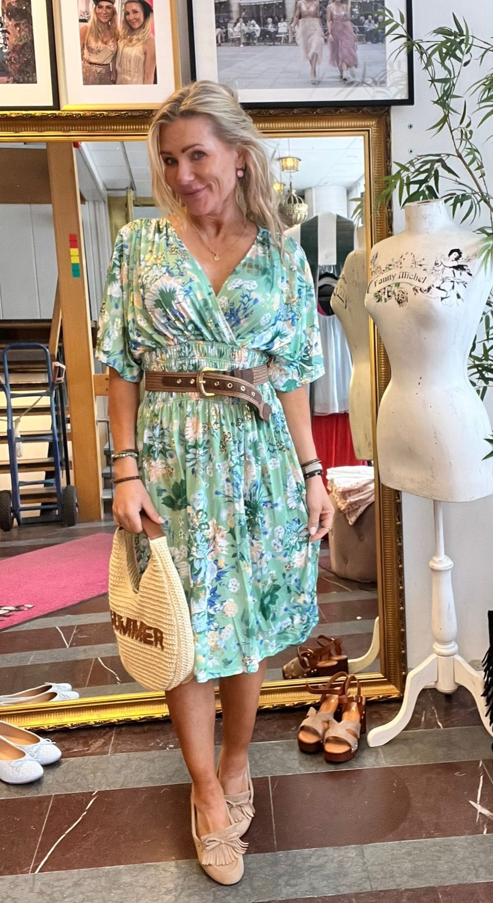 Blond kvinna poserar och ler i en butik, klädd i grön blommig klänning med brunt bälte och loafers, och håller en stråväska med texten “SUMMER”. Bakom syns guldspegel, skor, foton och provdocka med texten “Jenny Michel”.