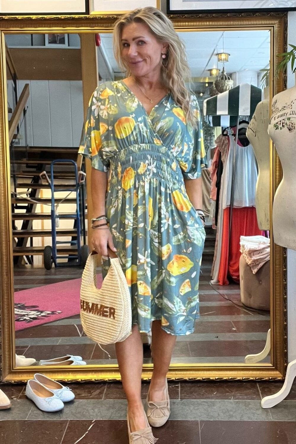 Leende kvinna står framför en stor guldramsspegel i en klädbutik och visar en blå klänning med citronmönster. Hon håller en stråväska med texten “SUMMER”, och skor samt klädställning syns i bakgrunden.