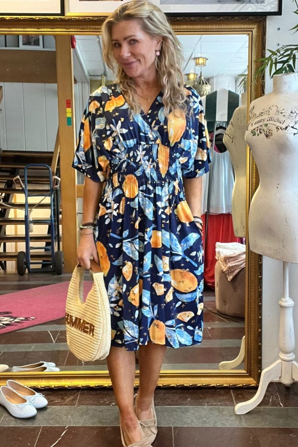 Blond kvinna poserar framför en stor guldramsspegel i en butik/studio, bär mörkblå klänning med frukt- och blommönster, beige loafers och stråväska med texten “TAMER”, omgiven av skyltdocka och inramade foton på väggen.