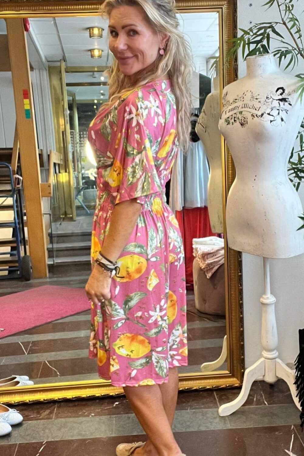 Kvinna i rosa klänning med citron- och blommönster står sidvänd och ler mot kameran i en butik. Hon provar outfit framför en stor guldramsspegel; intill syns en provdocka med texten ”Tommy Michel” och en teckning.
