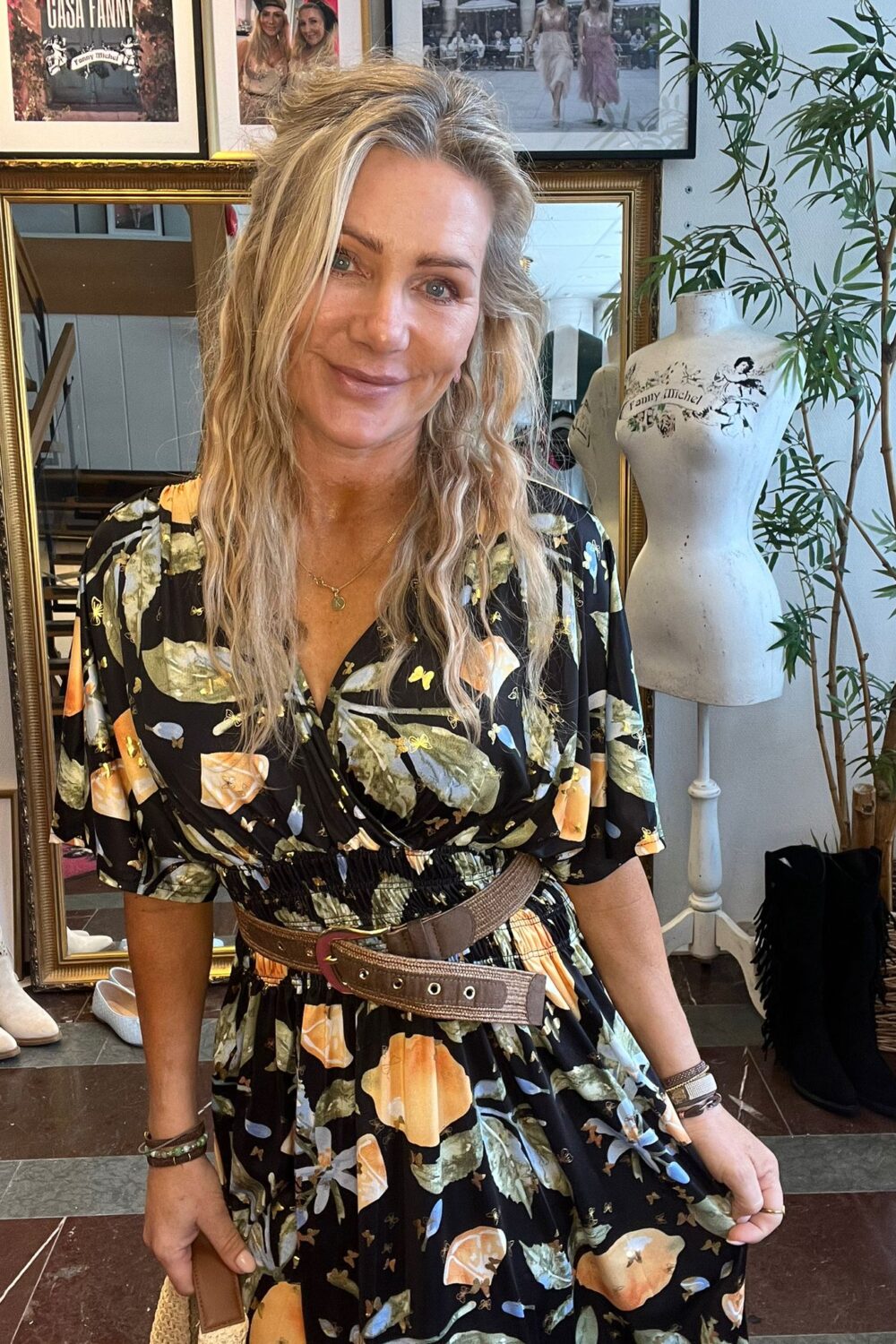 Blond kvinna står och ler mot kameran i en butik/ateljé, klädd i svart blommig klänning med brett bälte. Bakom syns en stor spegel, inramade foton, en provdocka med texten “Fanny Michel” och en hög växt.