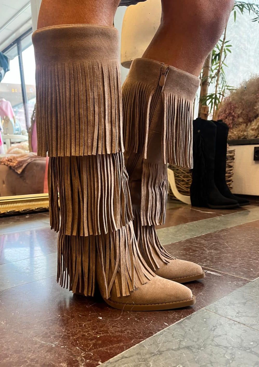 En person står inomhus och visar bruna, knähöga mockastövlar med flera lager långa fransar och dragkedja på sidan. Stövlarna står på marmorgolv i en butiksmiljö med andra skor i bakgrunden.