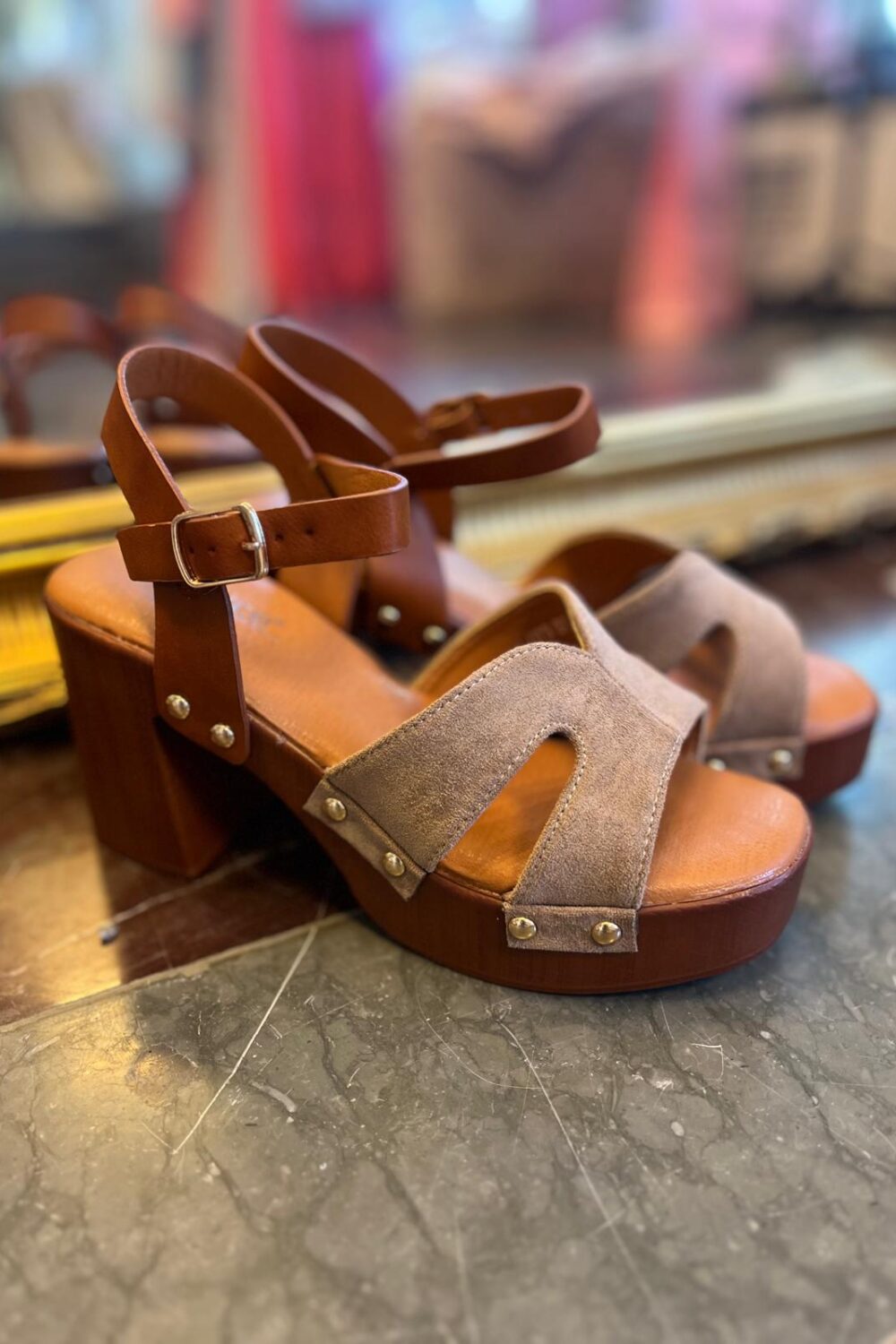 Bruna platåsandaler med klack står på ett grått marmorgolv, med läderremmar och spänne samt ljusbruna mockadetaljer med nitar. Skorna är i fokus medan butiksmiljön i bakgrunden är suddig.
