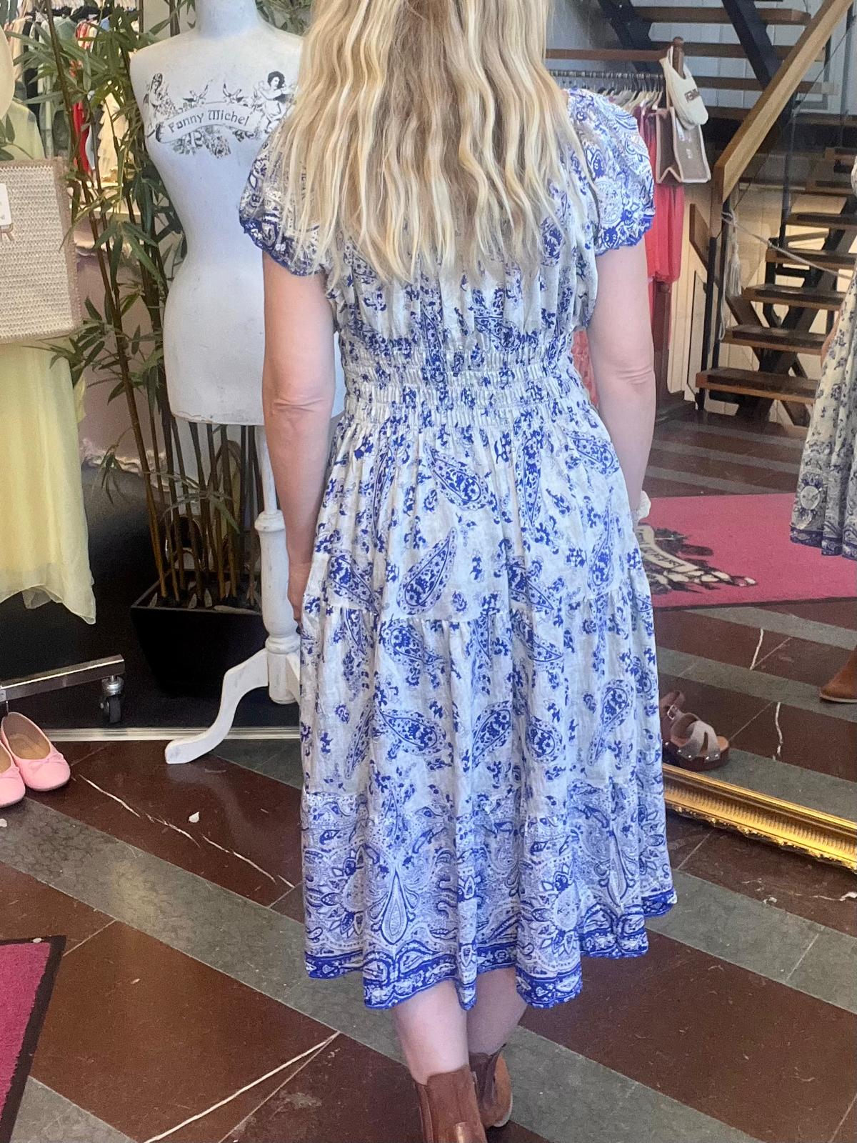 Kvinna med långt blont hår står med ryggen mot kameran i en butik och visar en vit, blåmönstrad klänning med rynkad midja och puffärmar. Bredvid syns en provdocka med texten “Fanny Michel” och klädställningar.