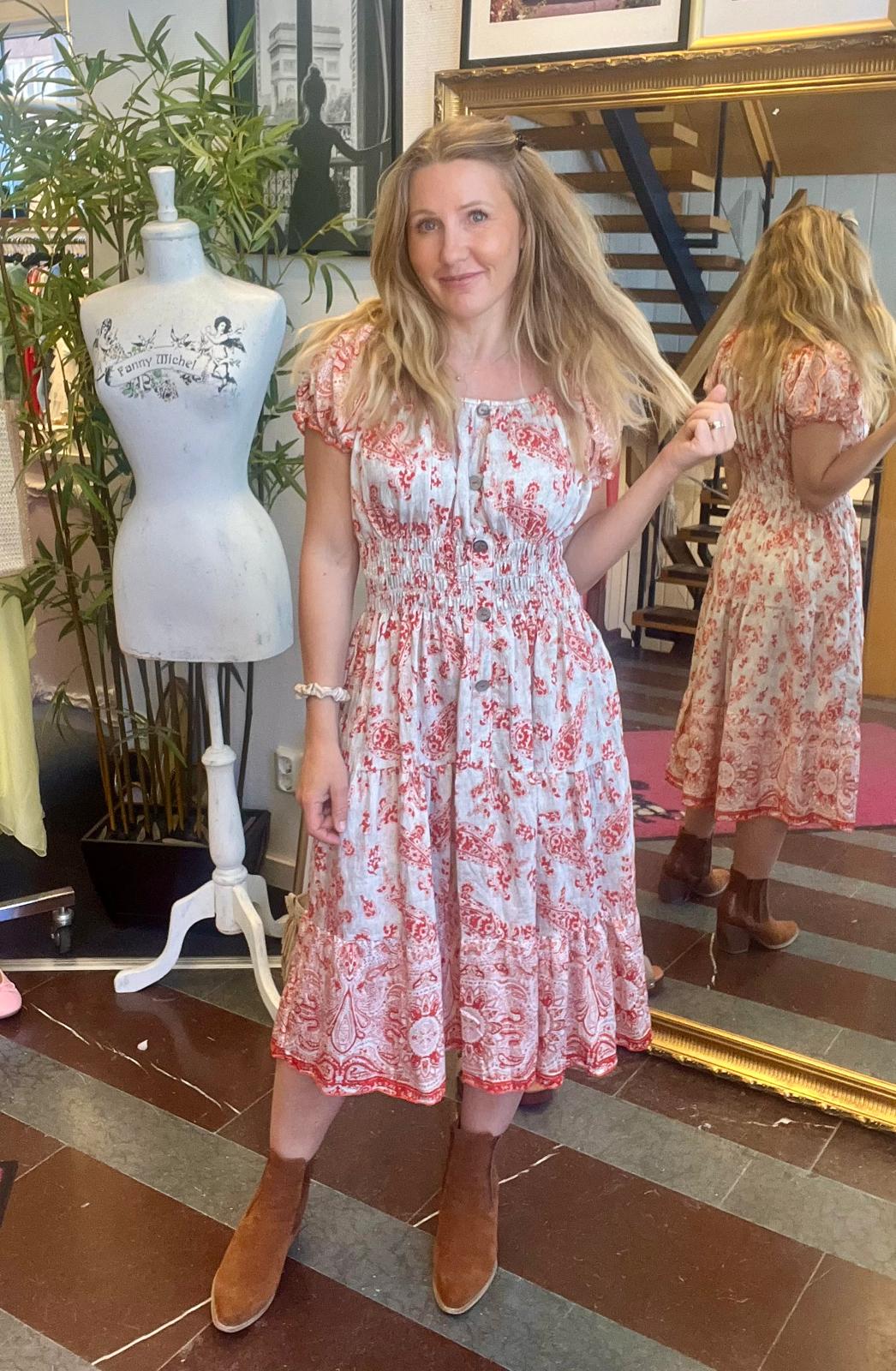 Kvinna i rödvit mönstrad klänning står i en butik och håller upp en hårslinga medan hon ler mot kameran. Bredvid finns en provdocka med texten “Fanny Michel” och en spegel som visar hennes rygg.