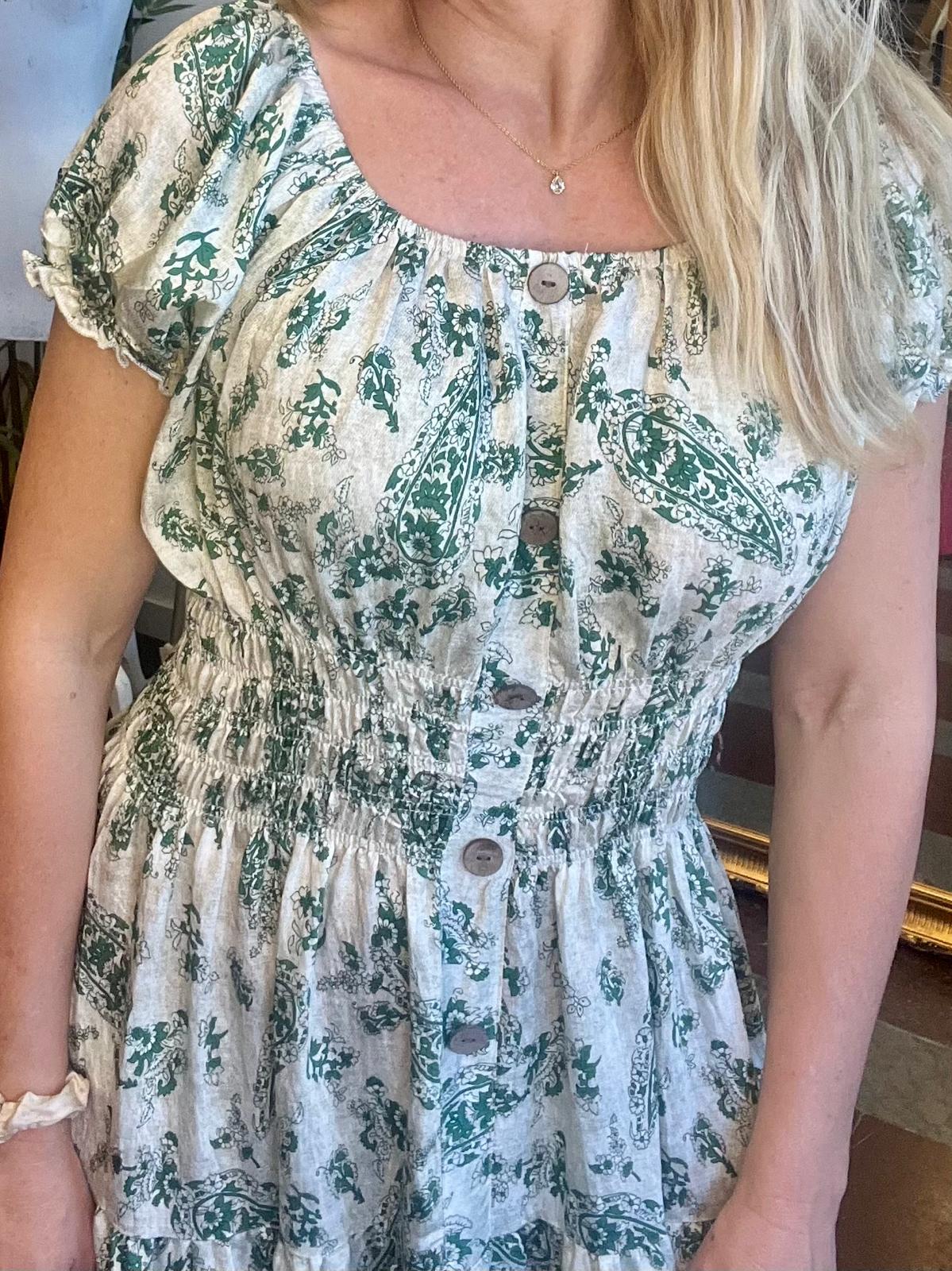 Kvinna står inomhus och visar en ljus klänning med grönt blommigt mönster, knappslå framtill och rynkad midja. Hon har långt blont hår, ett halsband med liten sten och rynkade kortärmar.