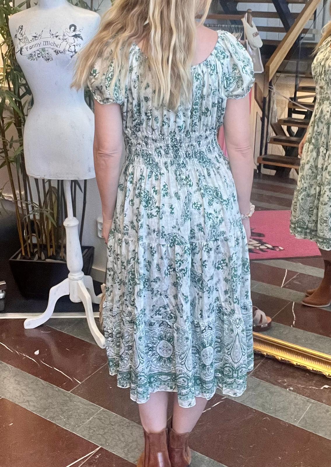 Kvinna står med ryggen mot kameran och provar en vit, grönmönstrad midiklänning med puffärmar och smock i midjan, framför en stor spegel i en butik. Till vänster syns provdocka med texten “Fanny Michel”.