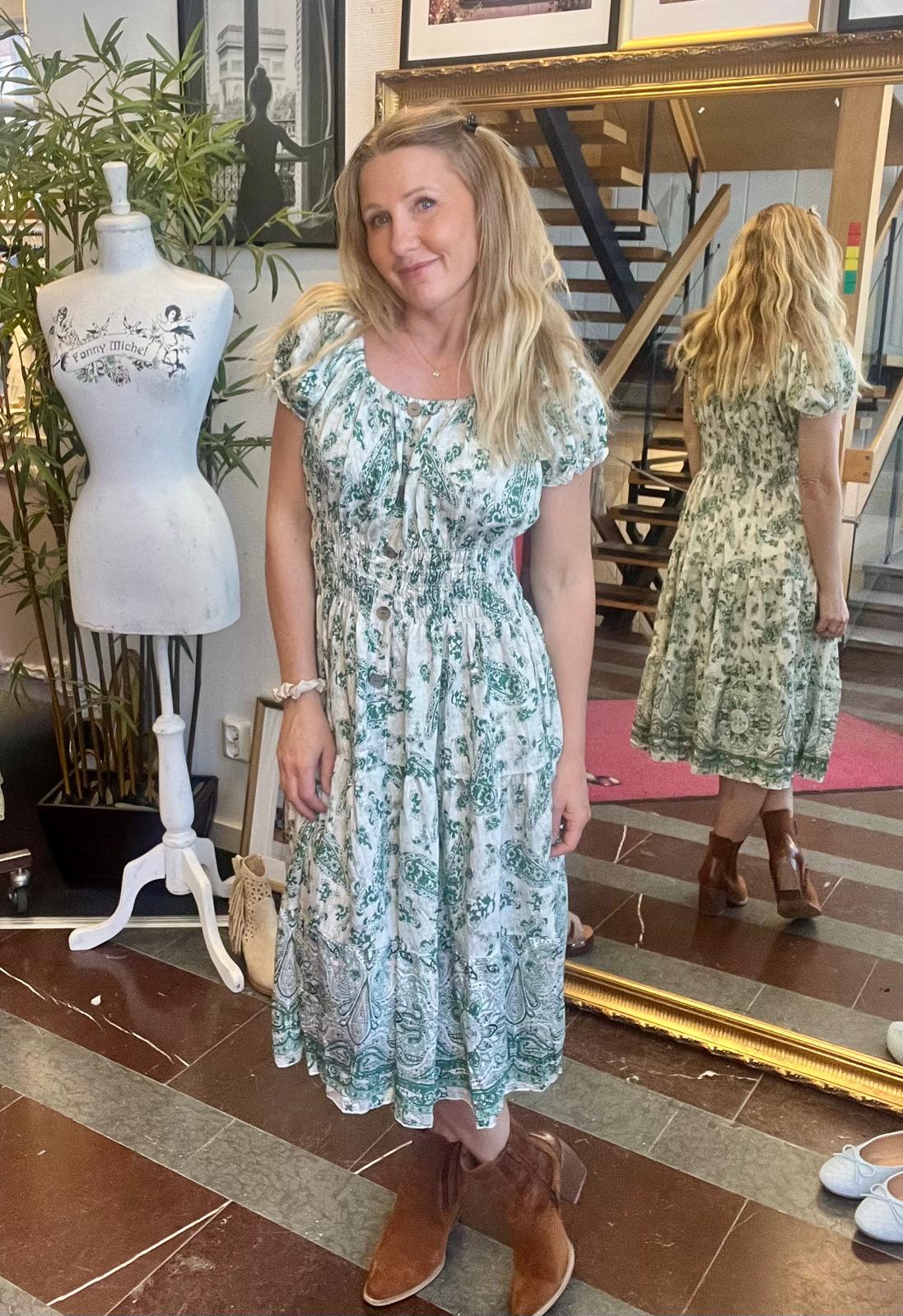 Kvinna står och poserar i en butik eller studio, iförd en vitgrön mönstrad klänning och bruna boots, med sin rygg speglad i en stor guldramsspegel. Bredvid syns provdocka med texten ”Fanny Michel”.
