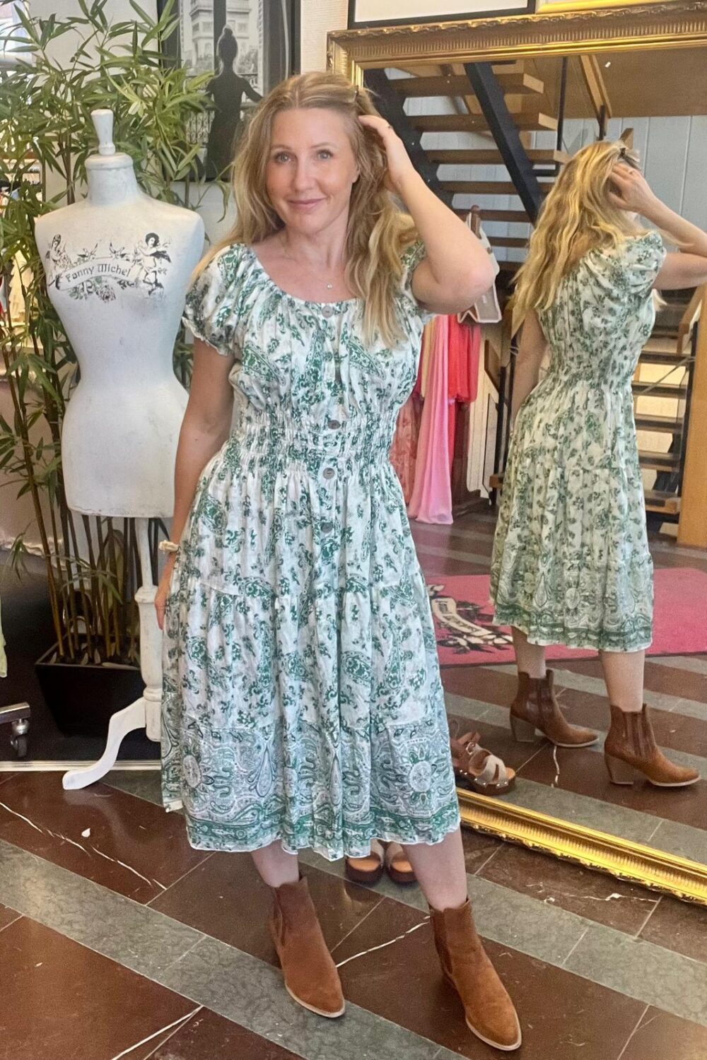 Kvinna i grönmönstrad klänning poserar och rör vid håret i en butik, med sin spegelbild bakom. Bredvid står en provdocka med texten “Fanny Michel”, och i bakgrunden syns trappa och hängande kläder.