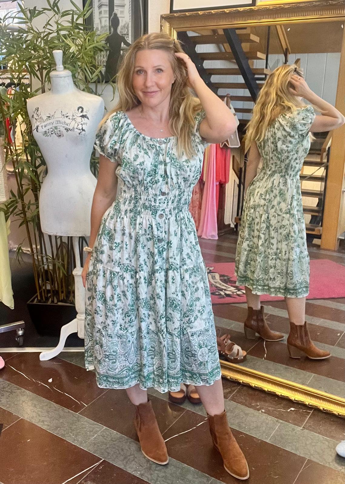 Kvinna i grönmönstrad klänning poserar och rör vid håret i en butik, med sin spegelbild bakom. Bredvid står en provdocka med texten “Fanny Michel”, och i bakgrunden syns trappa och hängande kläder.