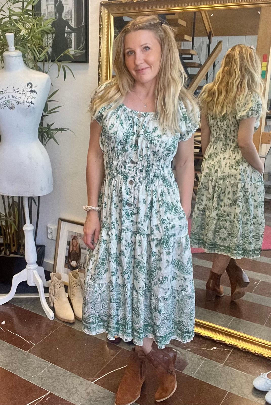 Kvinna står och poserar i en butik eller ateljé, iklädd en vitgrön mönstrad klänning och bruna boots. Hon speglas i en stor guldram och omges av provdocka, växter och skor på golvet.