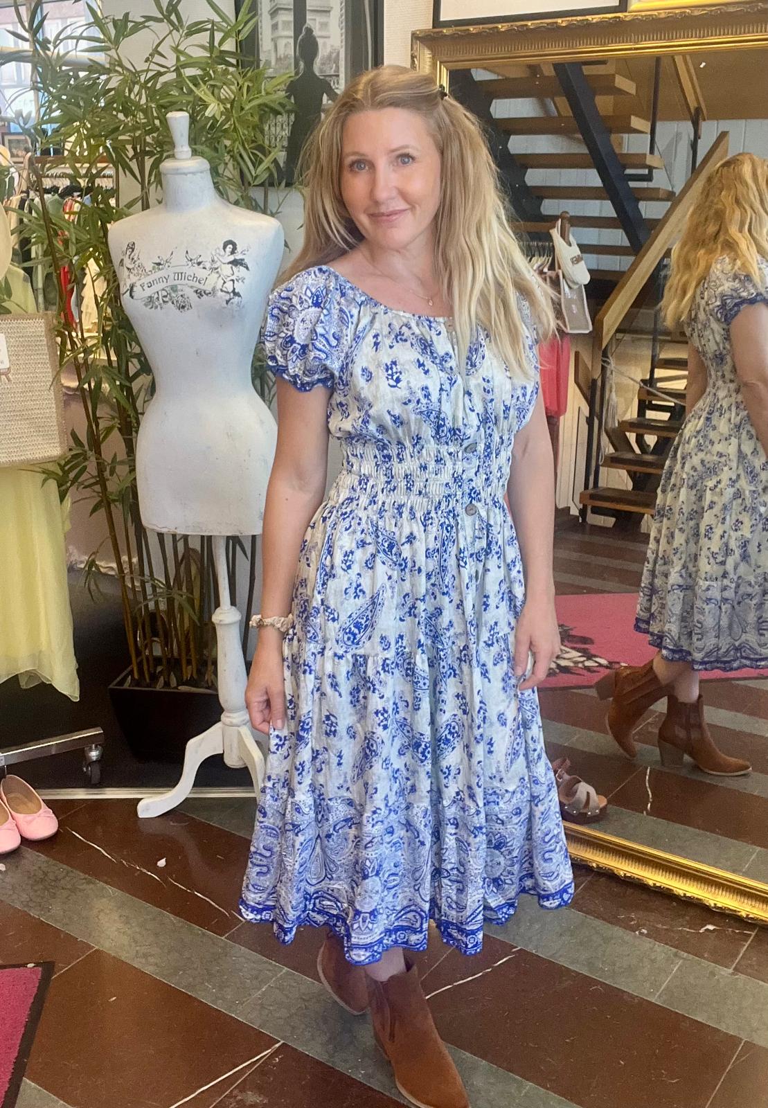 Kvinna står och poserar i en butik, iklädd blåvit mönstrad midiklänning och bruna boots. Bredvid finns en provdocka med texten “Fanny Michel”, och i en stor spegel syns hennes rygg samt en trappa och kläder.
