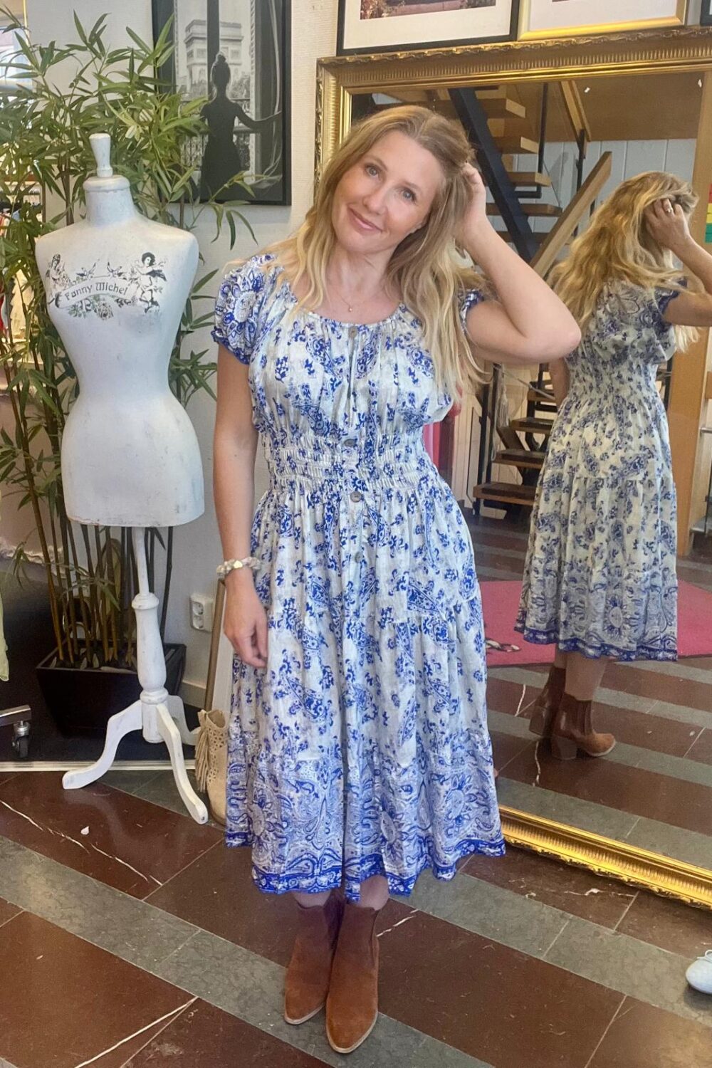 Kvinna i blå- och vitmönstrad midiklänning står och ler, med handen i håret, i en butik. Bakom syns hennes spegelbild och en provdocka med texten ”Fanny Michel”, samt trappa och växter.