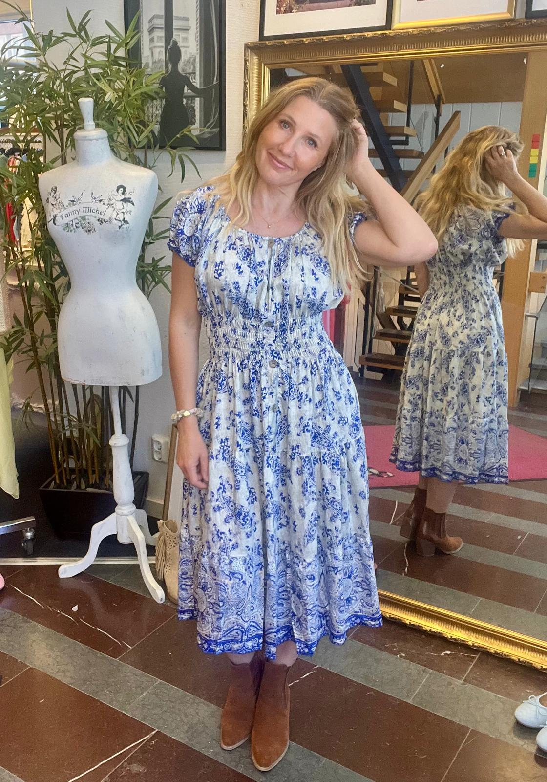 Kvinna i blå- och vitmönstrad midiklänning står och ler, med handen i håret, i en butik. Bakom syns hennes spegelbild och en provdocka med texten ”Fanny Michel”, samt trappa och växter.