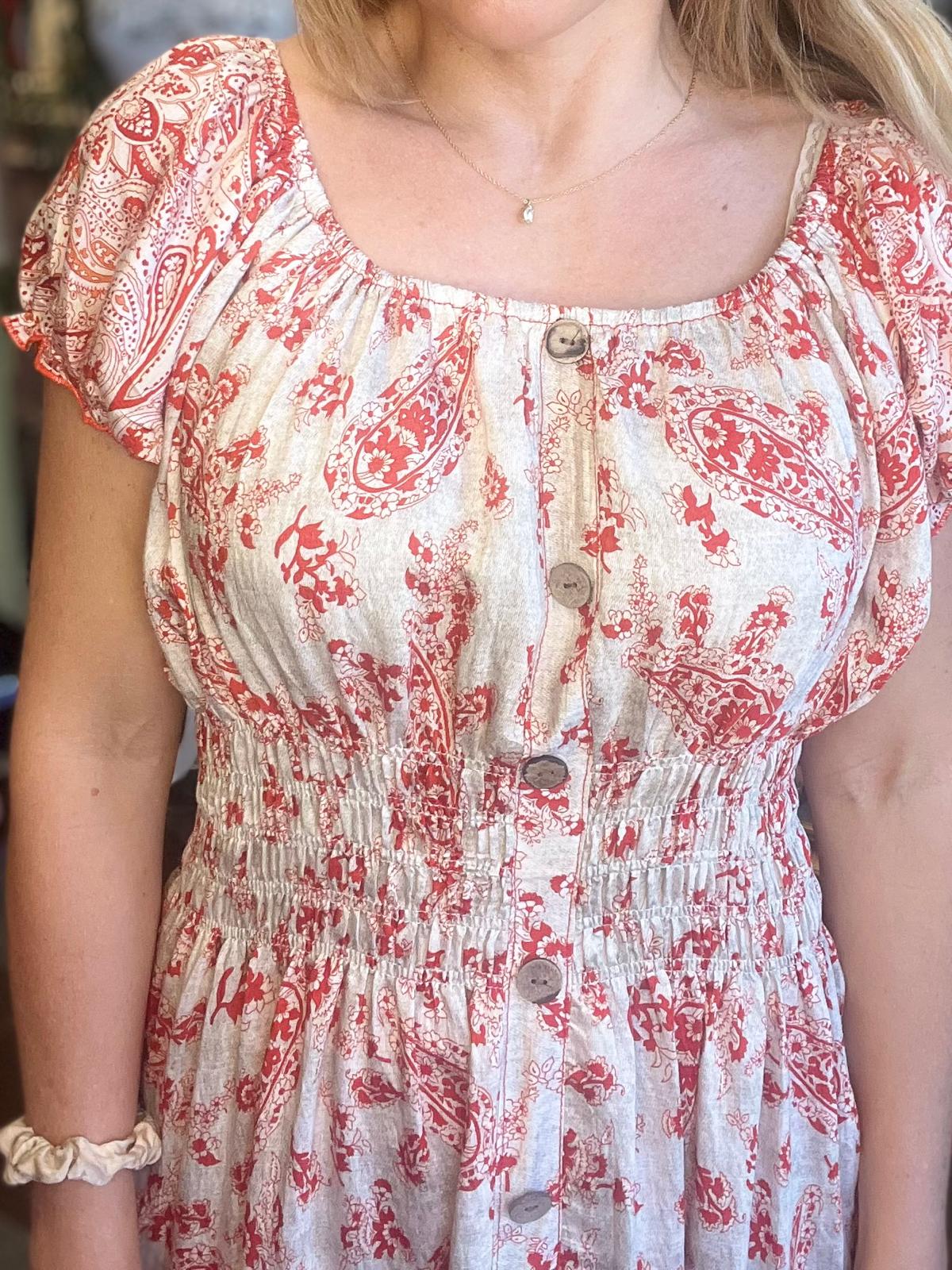 Person står vänd mot kameran och visar en ljus klänning med rött paisley- och blommönster. Klänningen har rundad urringning, korta ärmar, knappar framtill och resår i midjan; halsband och ljus scrunchie syns.