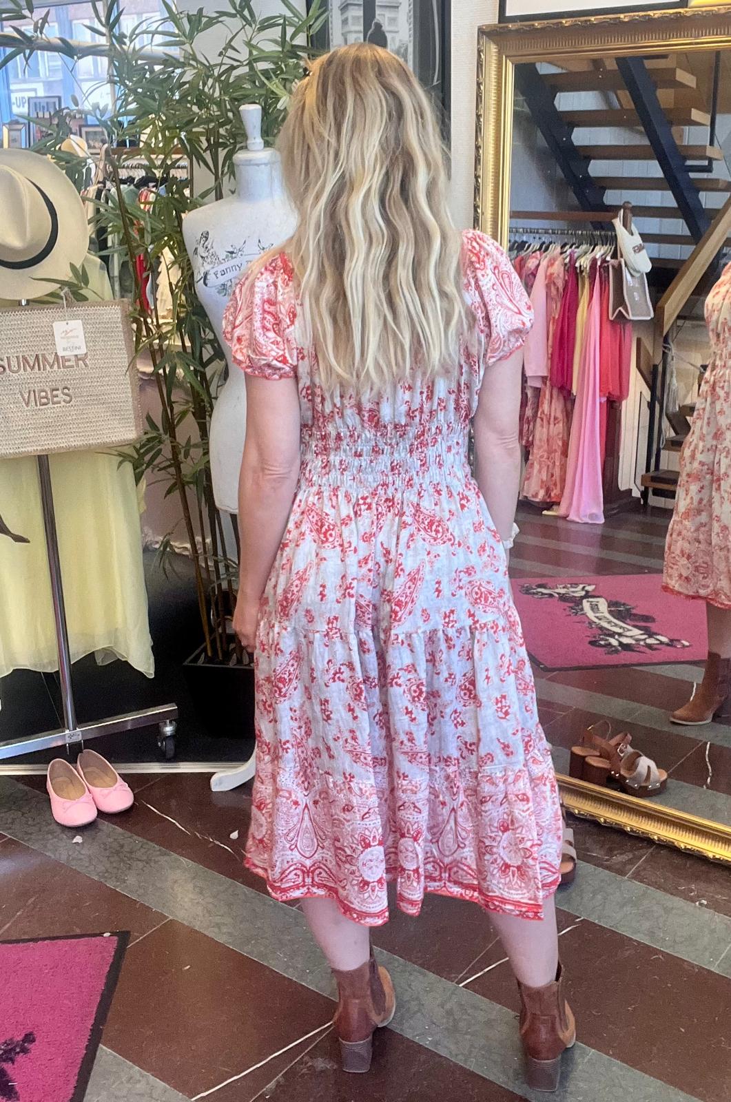 Kvinna med långt blont hår står med ryggen mot kameran och provar en vit klänning med rött mönster i en klädbutik. Bredvid syns skyltdocka, spegel, klädställning och skylttexten “SUMMER VIBES”.