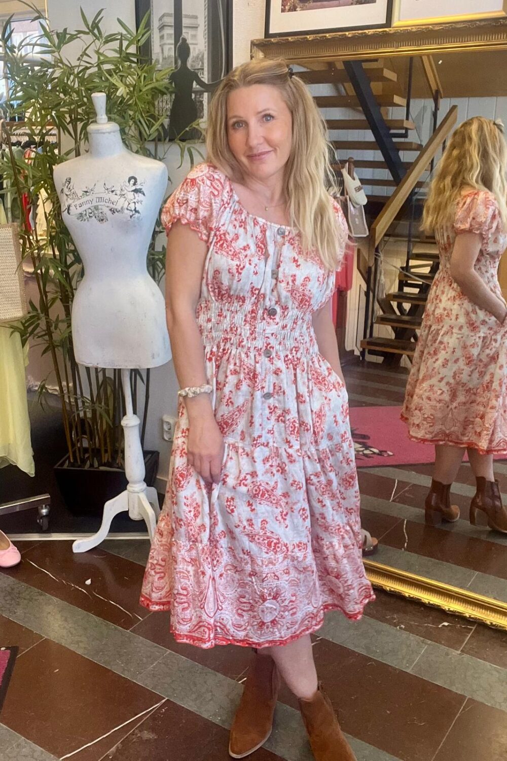 Kvinna står och poserar i en butik, iförd vit klänning med rött mönster och bruna boots, med händerna i fickorna. Bredvid syns en provdocka med texten “Fanny Michel” och en spegel visar hennes rygg.