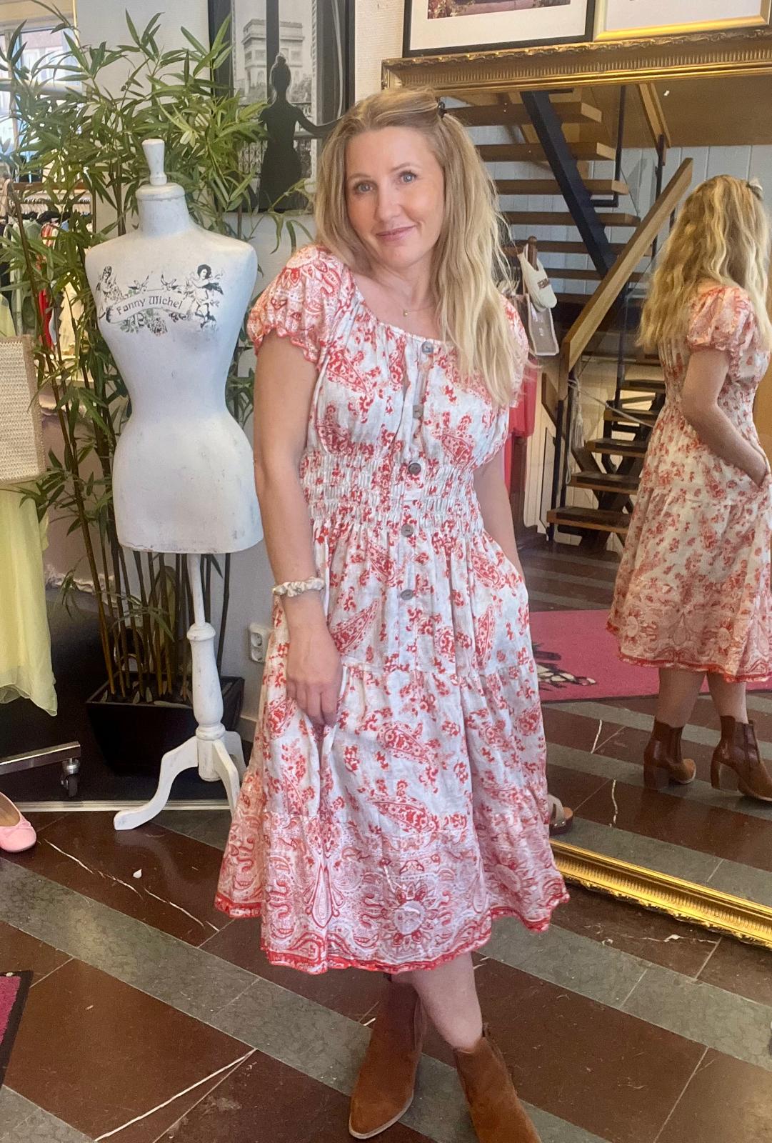 Kvinna står och poserar i en butik, iförd vit klänning med rött mönster och bruna boots, med händerna i fickorna. Bredvid syns en provdocka med texten “Fanny Michel” och en spegel visar hennes rygg.