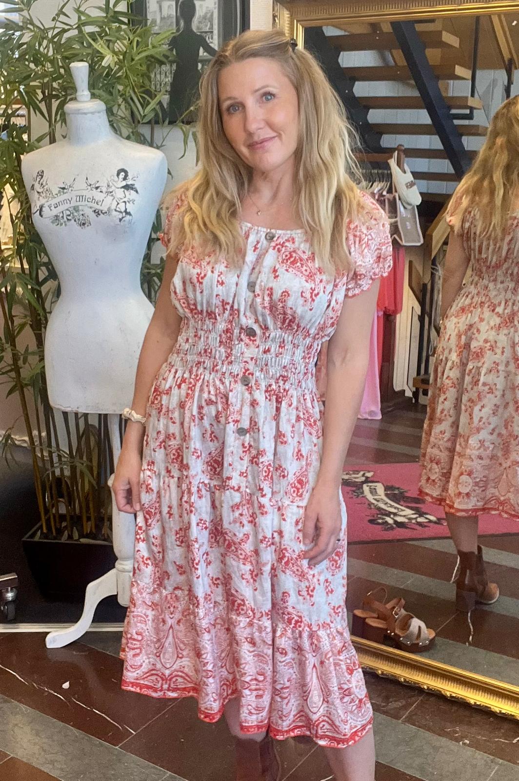 Kvinna står och poserar i en butik, iförd vit midiklänning med rött mönster och knappar framtill. Bredvid finns en provdocka med texten “Fanny Michel”, och en stor spegel visar hennes klänning bakifrån.