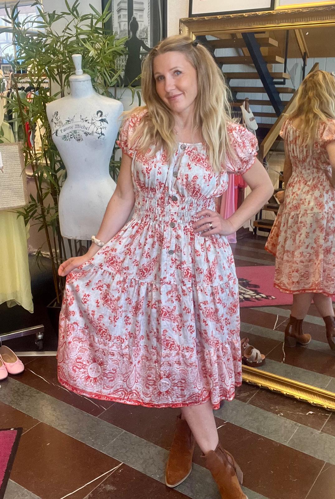 Kvinna i vit klänning med rött mönster poserar i en butik, håller ut kjolen och ler mot kameran. I bakgrunden syns provdocka med texten ”Fanny Michel” och hennes spegelbild bredvid en stor guldramsspegel.