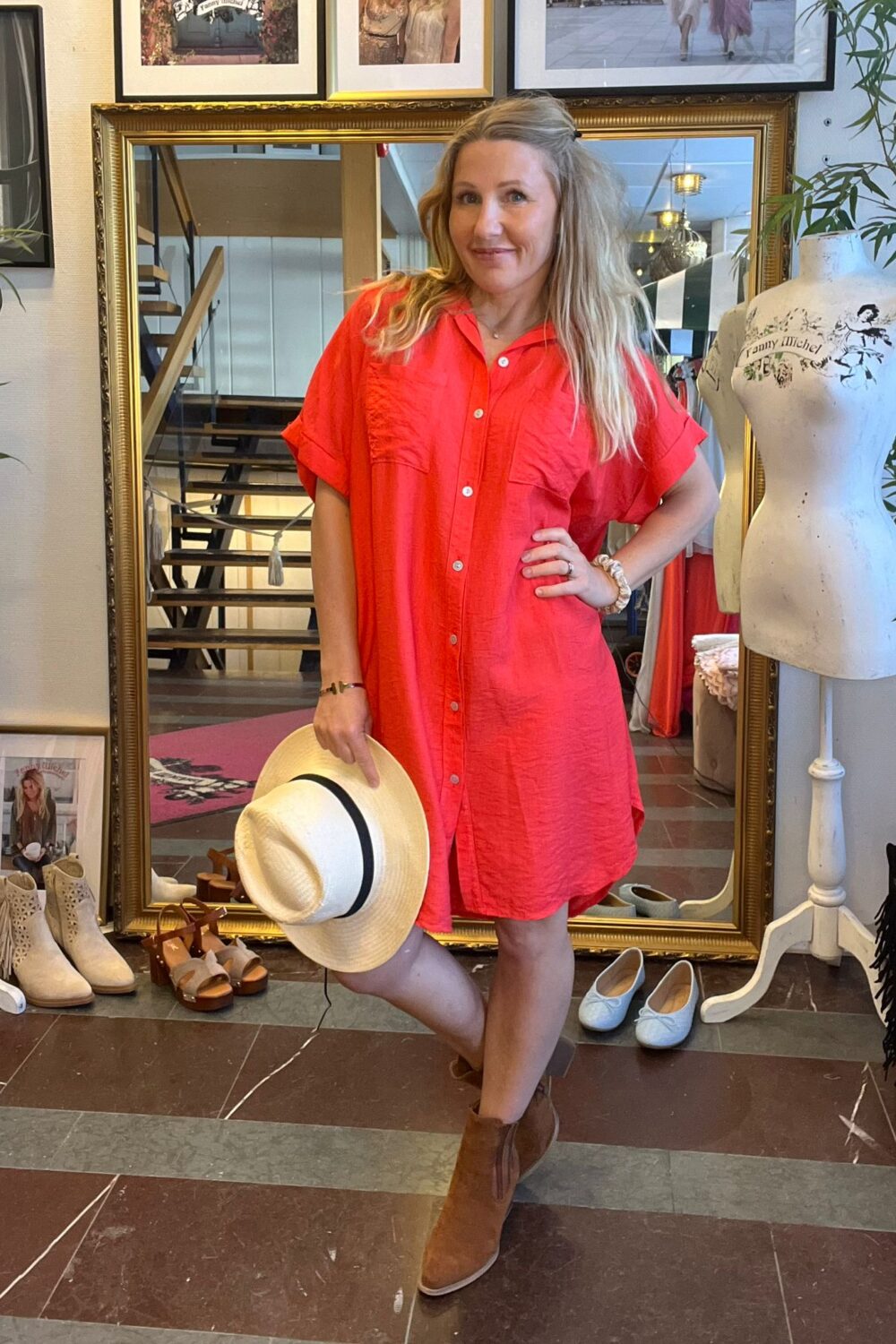 Kvinna i röd skjortklänning poserar med handen på höften och håller en ljus stråhatt med svart band. Hon står i en butik framför en stor guldspegel, omgiven av skor och skyltdocka.